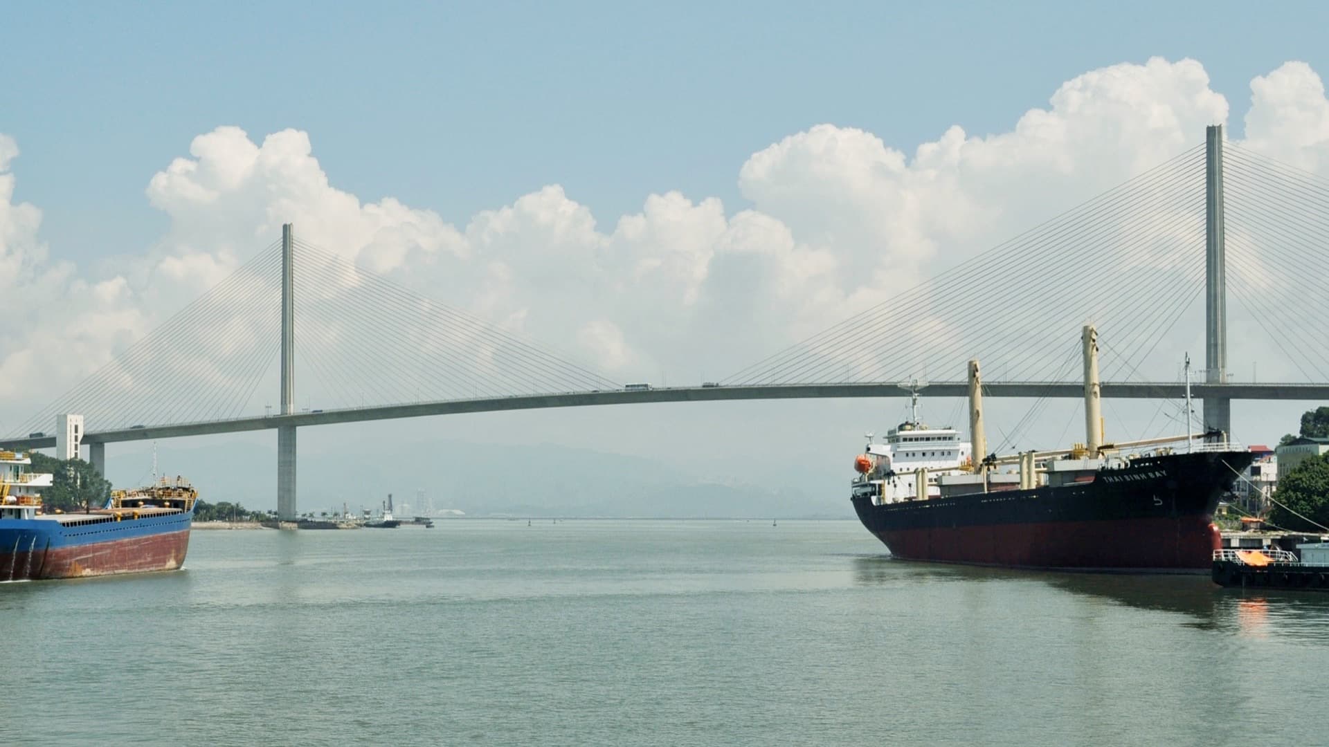 The cable-stayed Bai Chay Bridge spans the water at Ha Long under billowing white clouds, with cargo ships in the foreground.