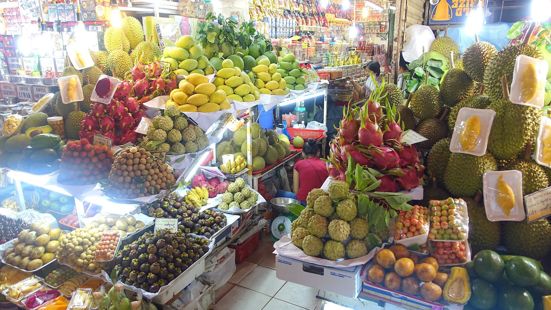 Fruit stalls packed with durian, dragon fruit, mangoes, mangosteens and custard apples inside Ben Thanh Market in Ho Chi Minh City, with vendors at work and handwritten price boards propped among the produce