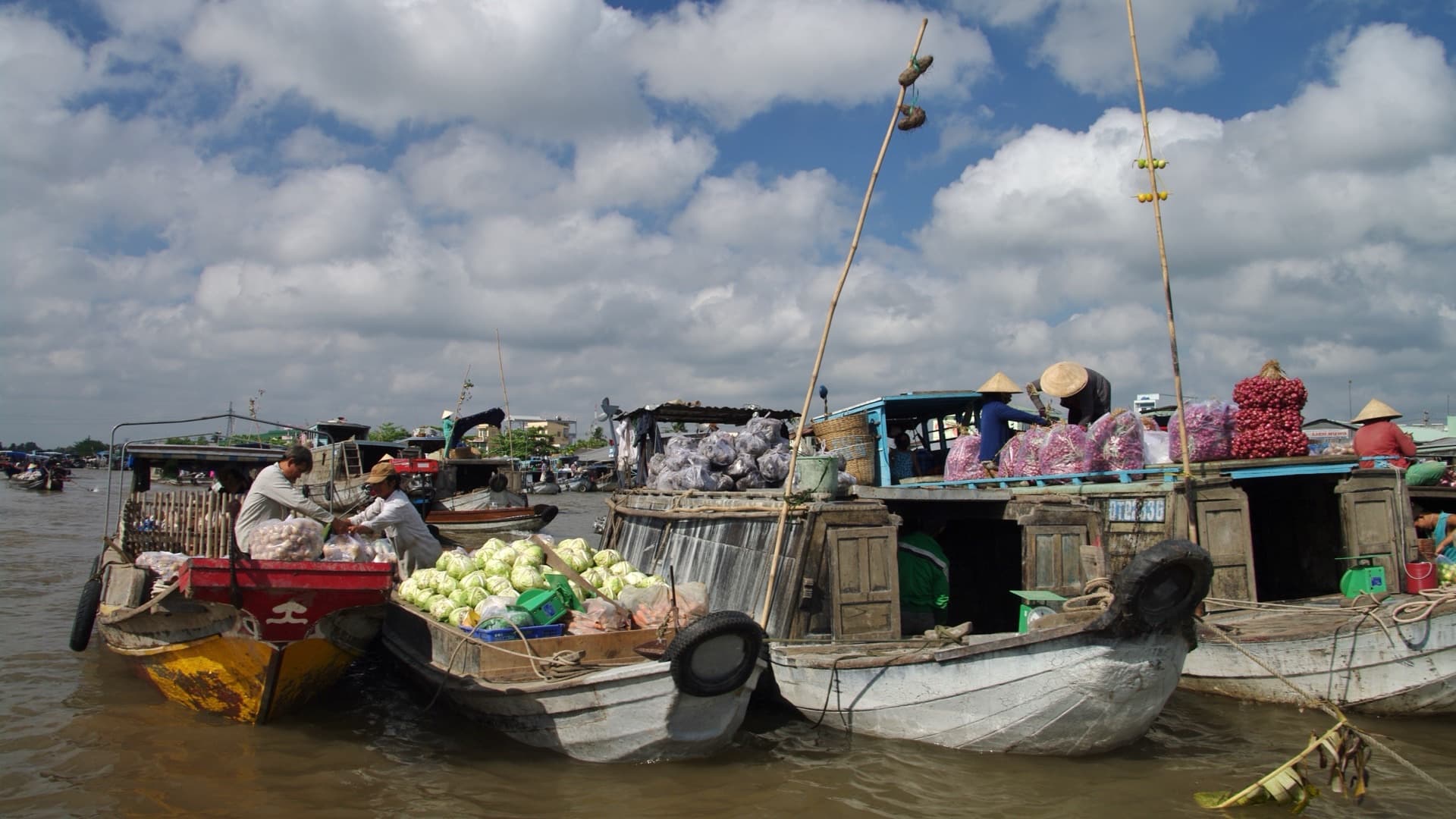 Wooden trading boats rafted together at Cai Rang floating market outside Can Tho, decks stacked with cabbages, garlic and sacks of red onions under a bank of cumulus cloud