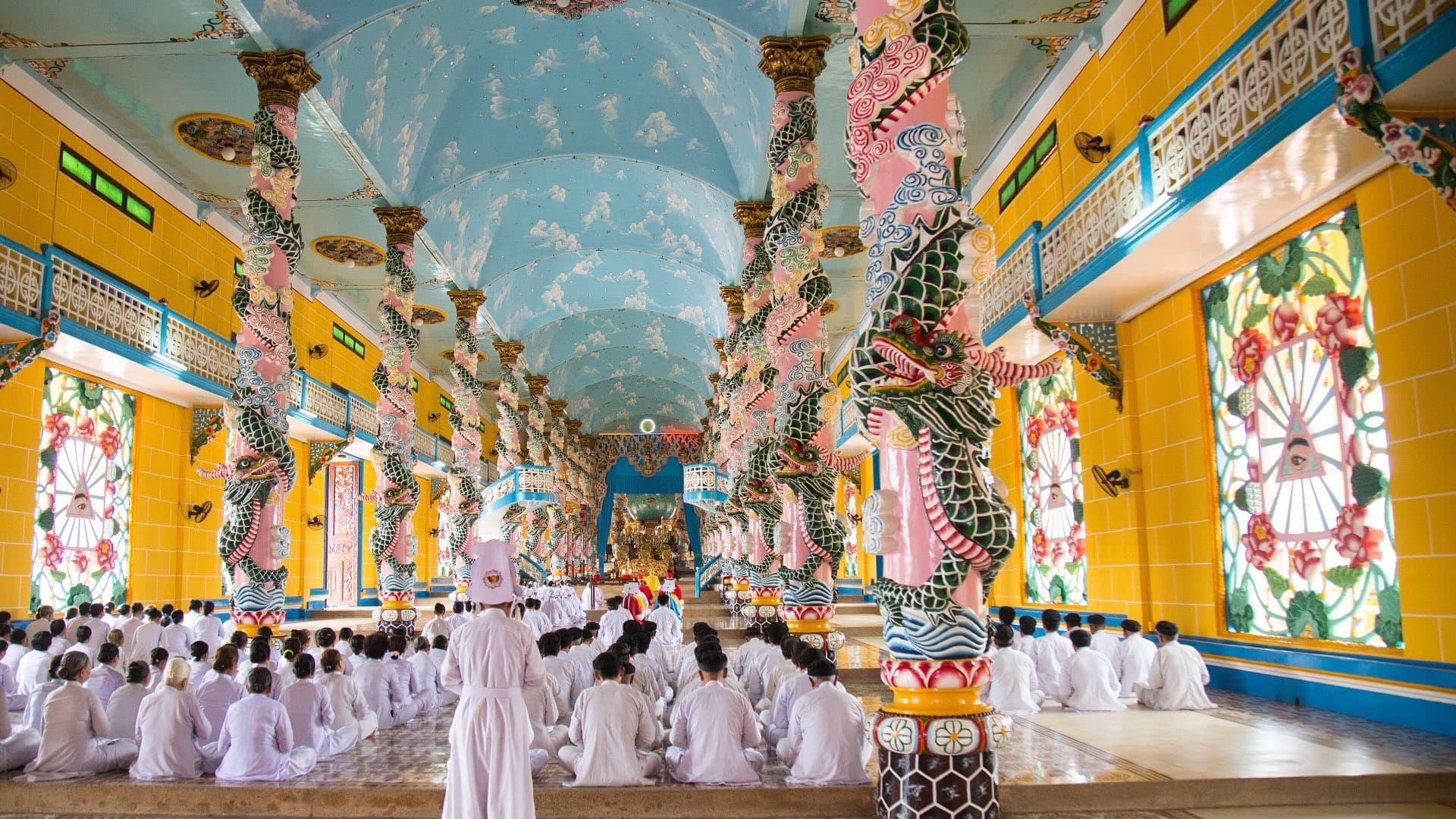 White-robed worshippers sit in rows beneath the sky-blue vaulted ceiling and dragon-wrapped columns inside the Cao Dai Holy See in Tay Ninh.