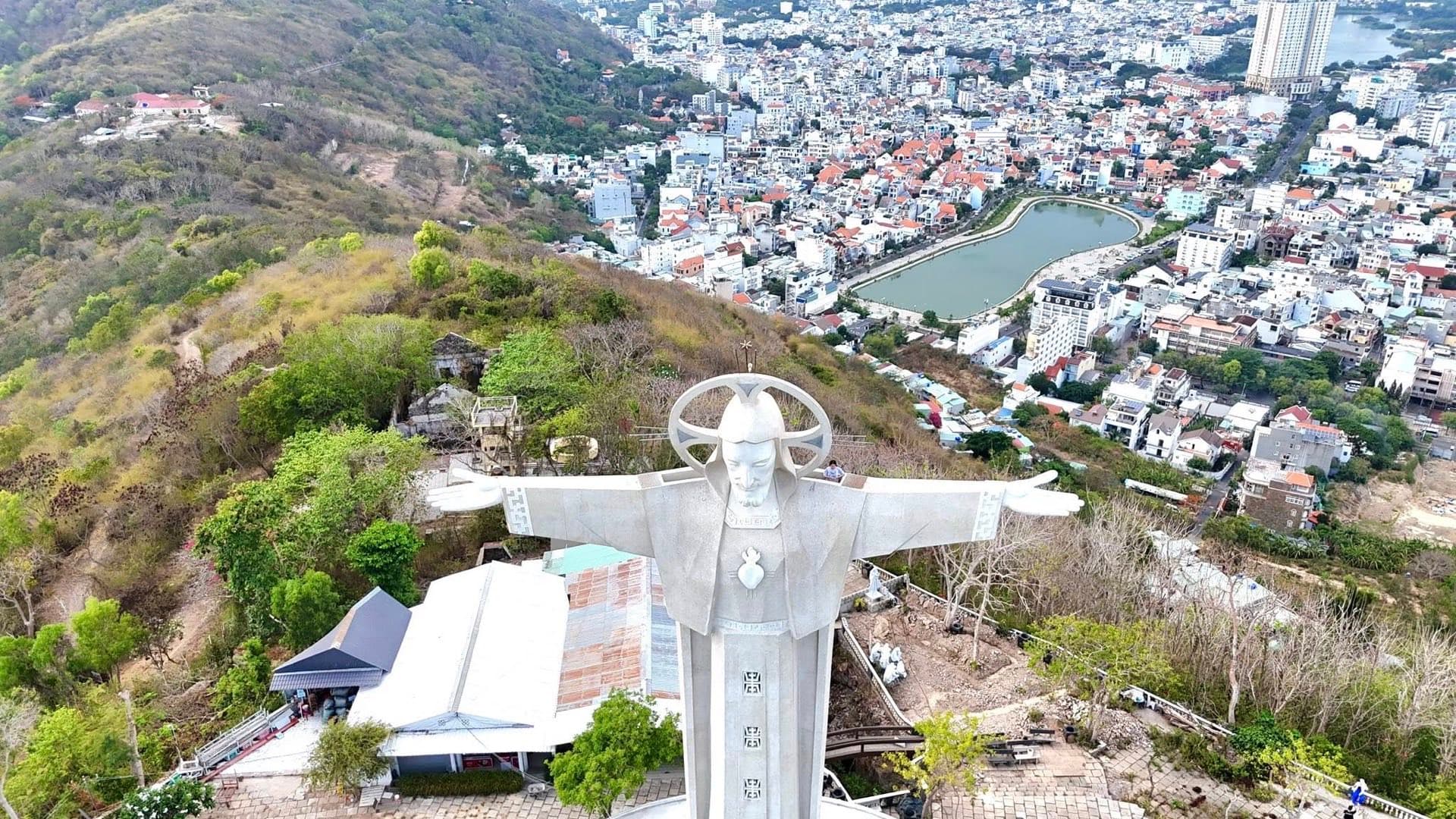 Aerial view of the white Christ of Vung Tau statue with outstretched arms on its hillside, overlooking the dense rooftops and a lake in Vung Tau city.