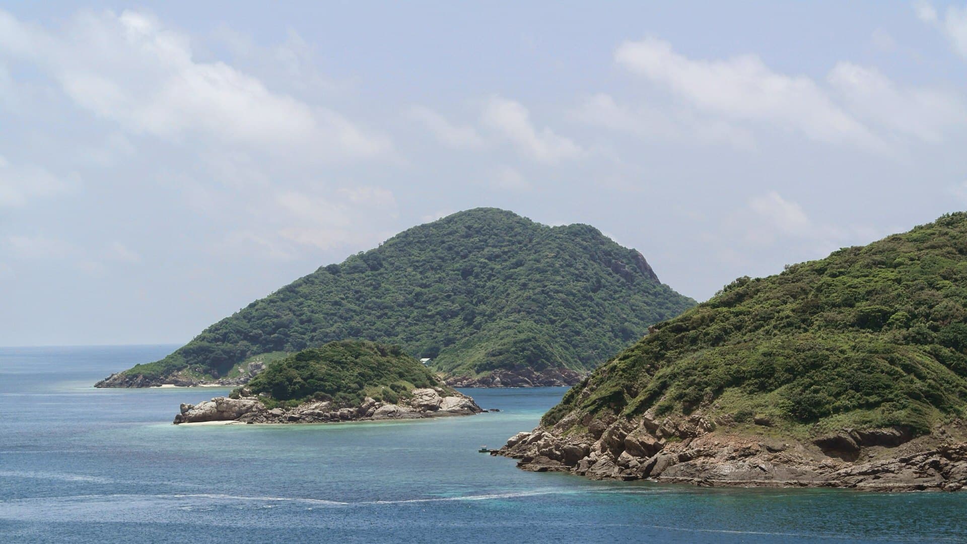 Forested green islets with rocky shores rise from blue sea off Ben Dam in Con Dao beneath a sky of soft white clouds.