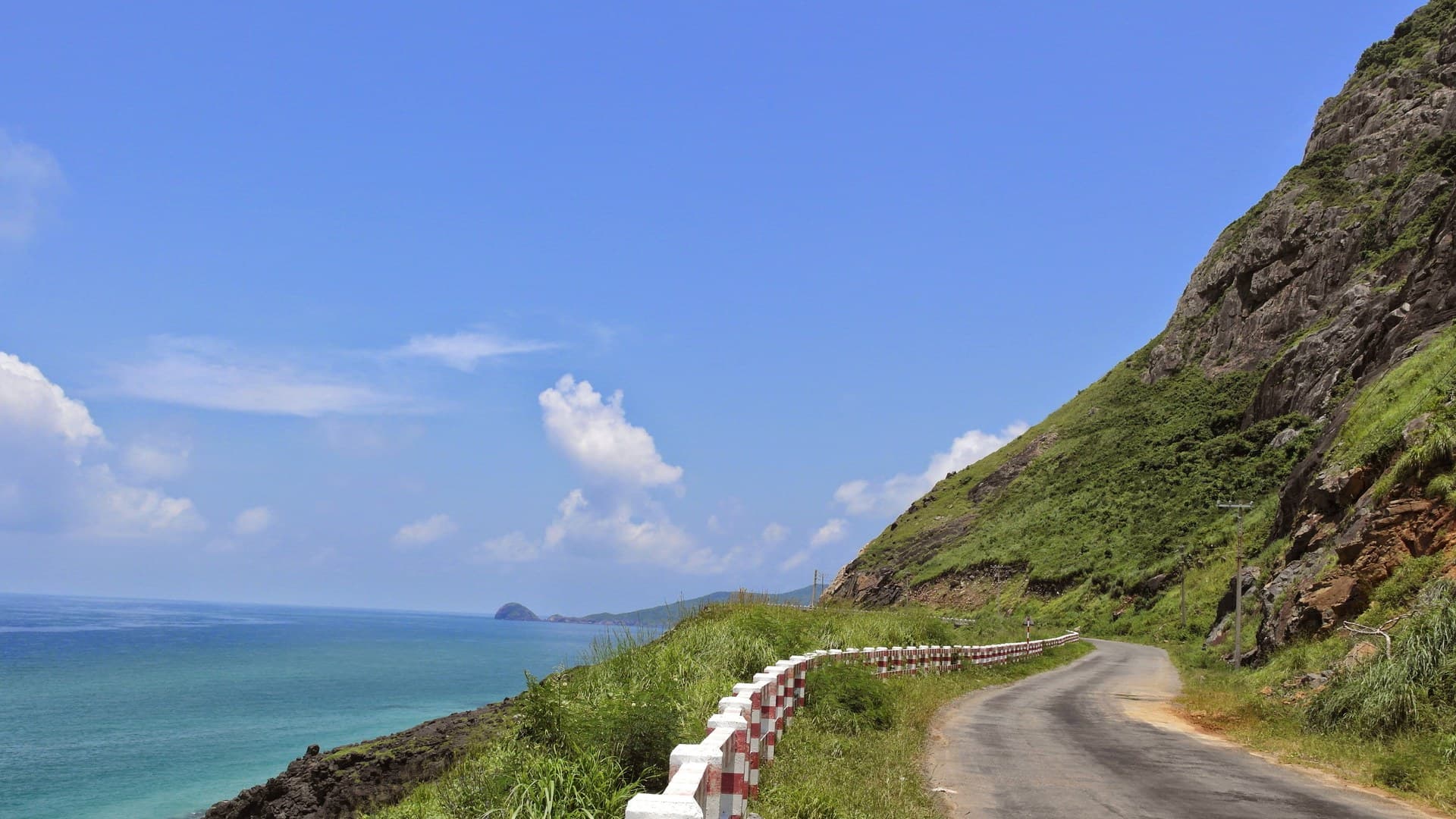 The coast road curving along a steep green headland on Con Son Island in Con Dao, above clear turquoise water with a small rocky islet offshore