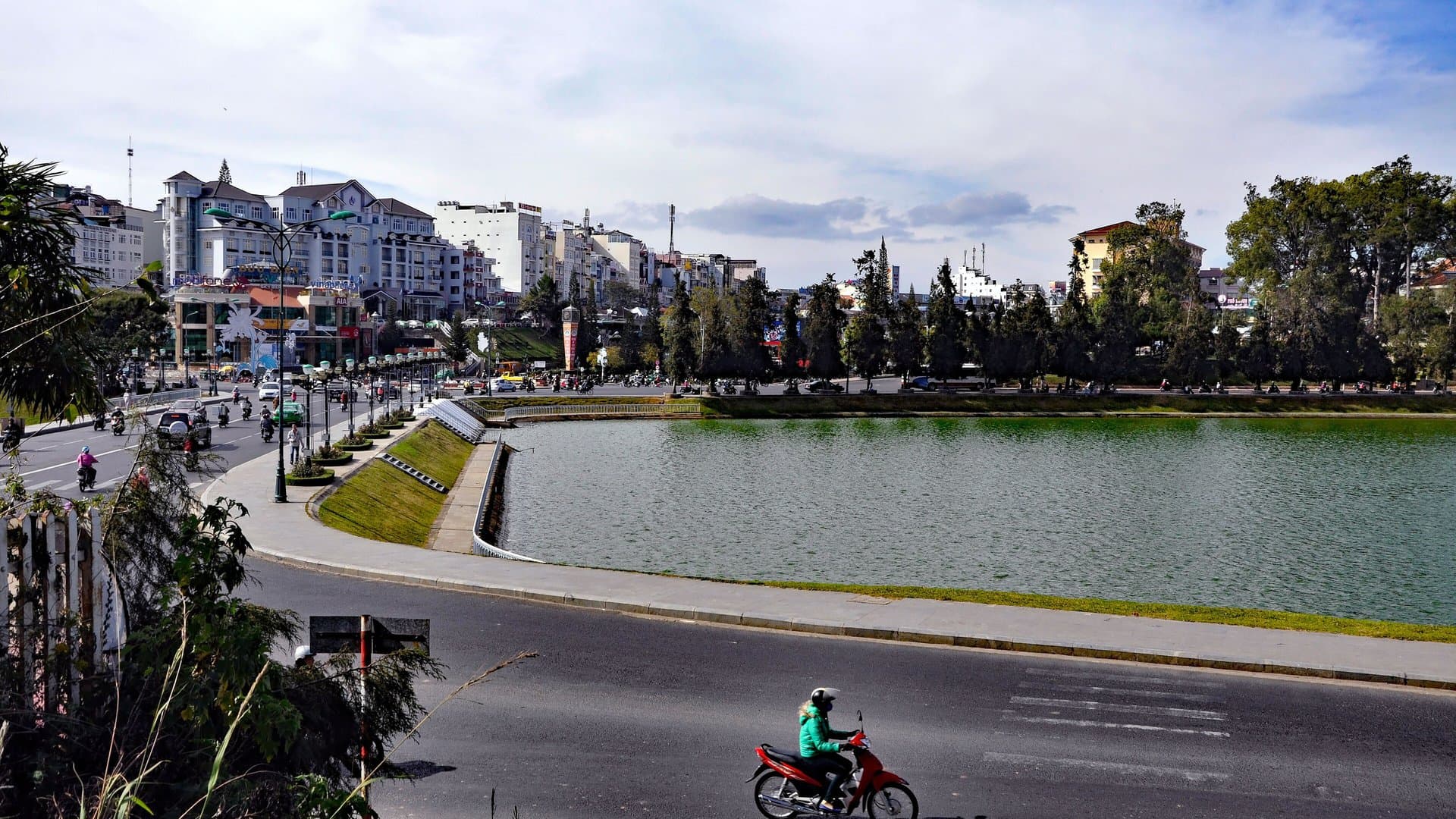 Xuan Huong Lake in central Da Lat under a blue sky with white clouds, with hotels and pine trees along the shore and a motorbike on the lakeside road