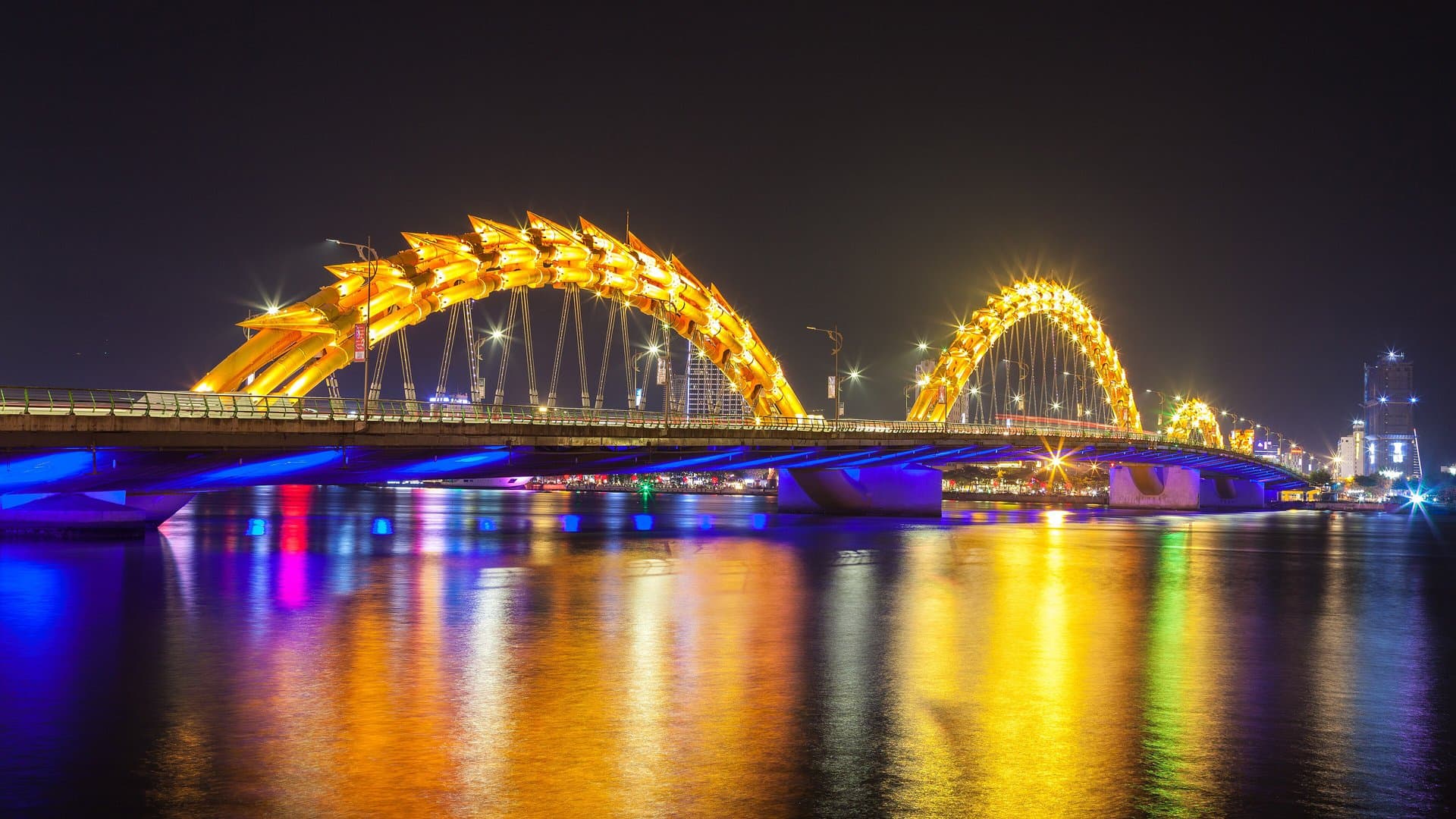 Da Nang's Dragon Bridge lit gold, blue and violet at night, its reflection stretching across the Han River