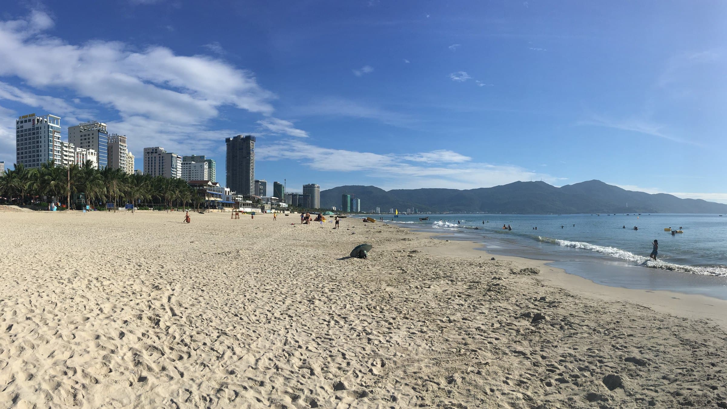 Summer morning on My Khe beach in Da Nang — swimmers in the shallows, hotel towers behind the coconut palms, and the Son Tra range across the bay