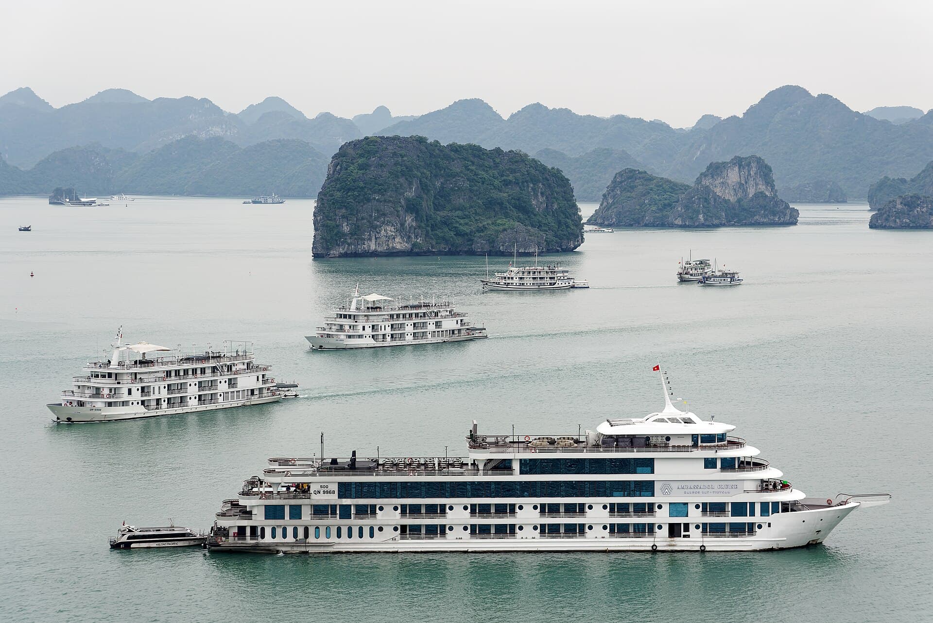 Multiple large cruise ships clustered around a single Ha Long Bay karst island — the visual proxy for the overtourism pressure documented in the 2025 Hoa Binh University study