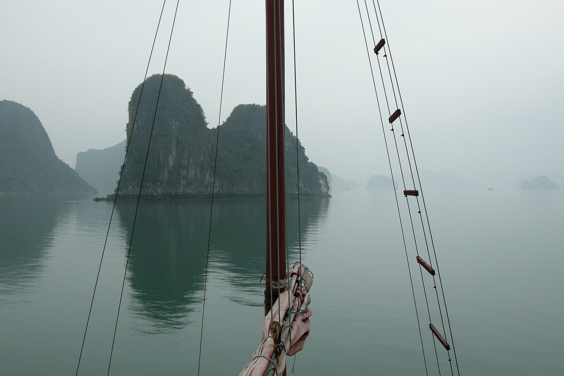 View through the rigging of a sailing junk in Ha Long Bay, mist-shrouded karst islands reflected in calm water — the iconic day-trip vantage point