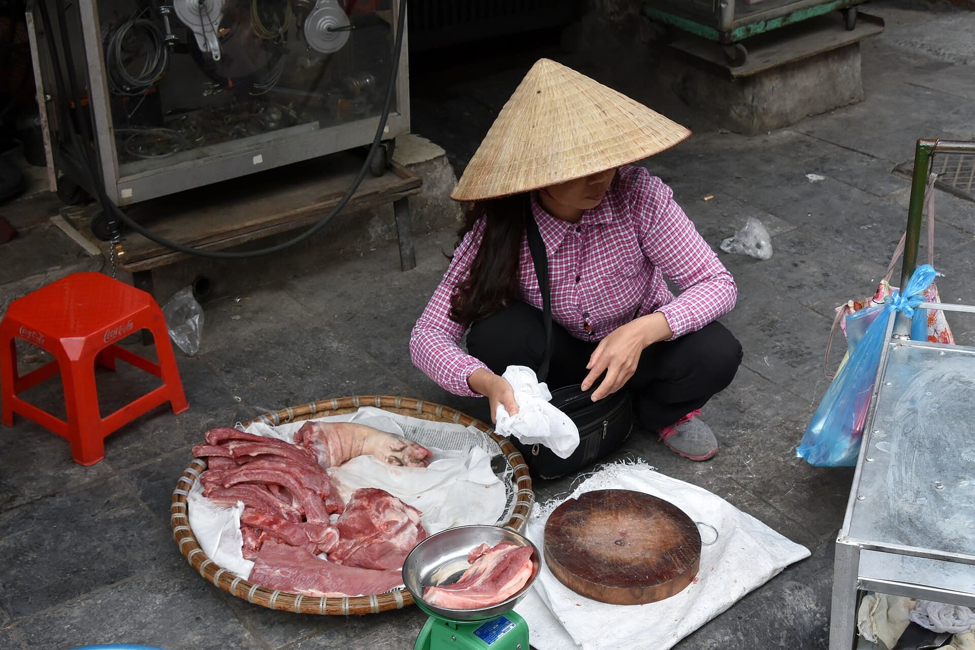 A Hanoi Old Quarter market vendor in conical hat preparing pork at her sidewalk stall — the everyday Vietnamese street life solo female travelers actually navigate