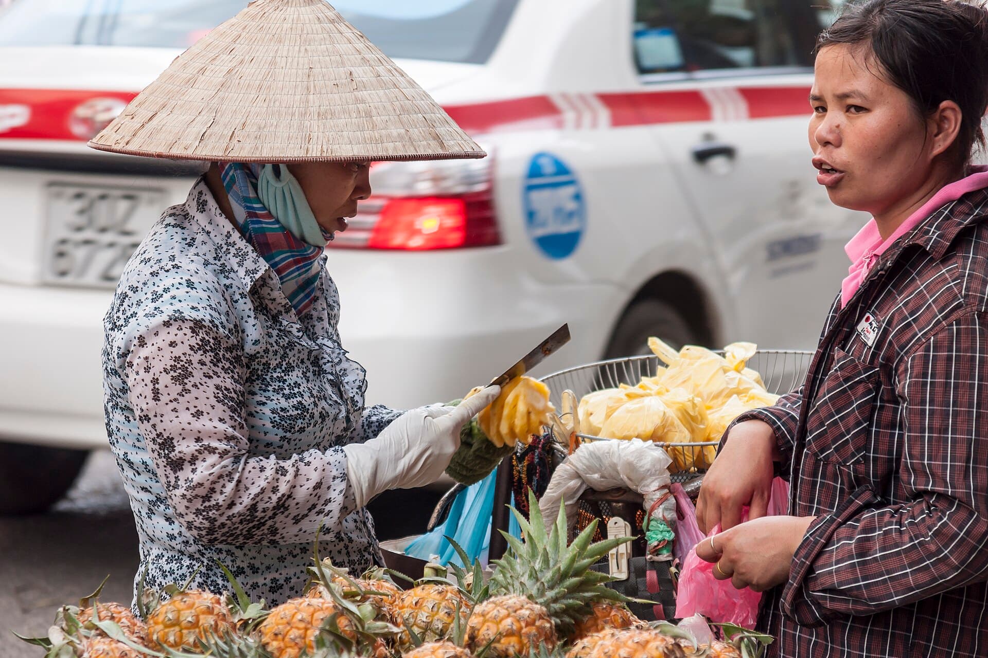 A pineapple street vendor in Hanoi handing fruit to a customer near a taxi — the everyday street commerce where most Vietnam hidden-cost decisions are made