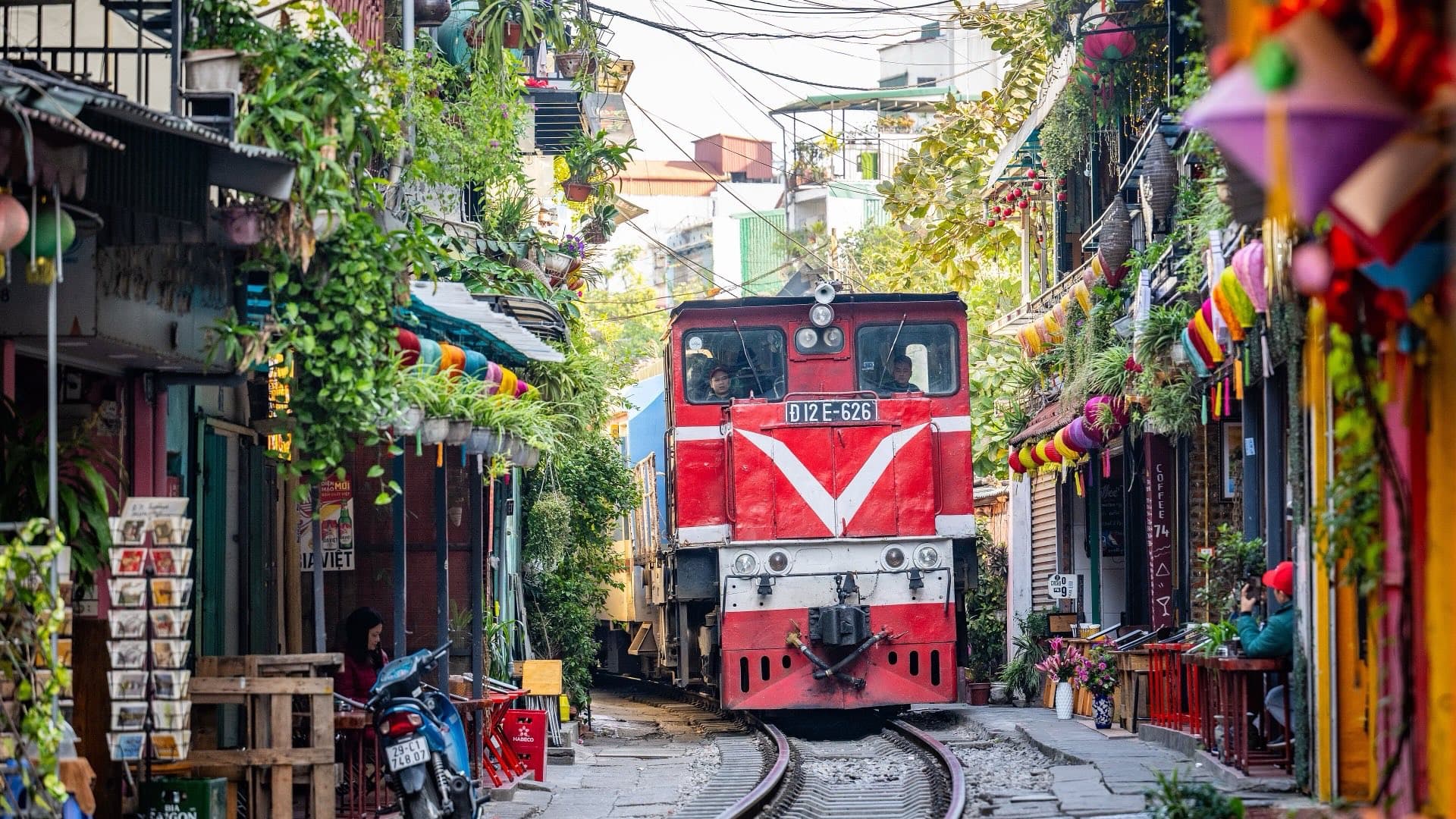 A red diesel locomotive edges along the narrow track of Hanoi's Train Street between plant-covered houses, cafes and hanging lanterns.
