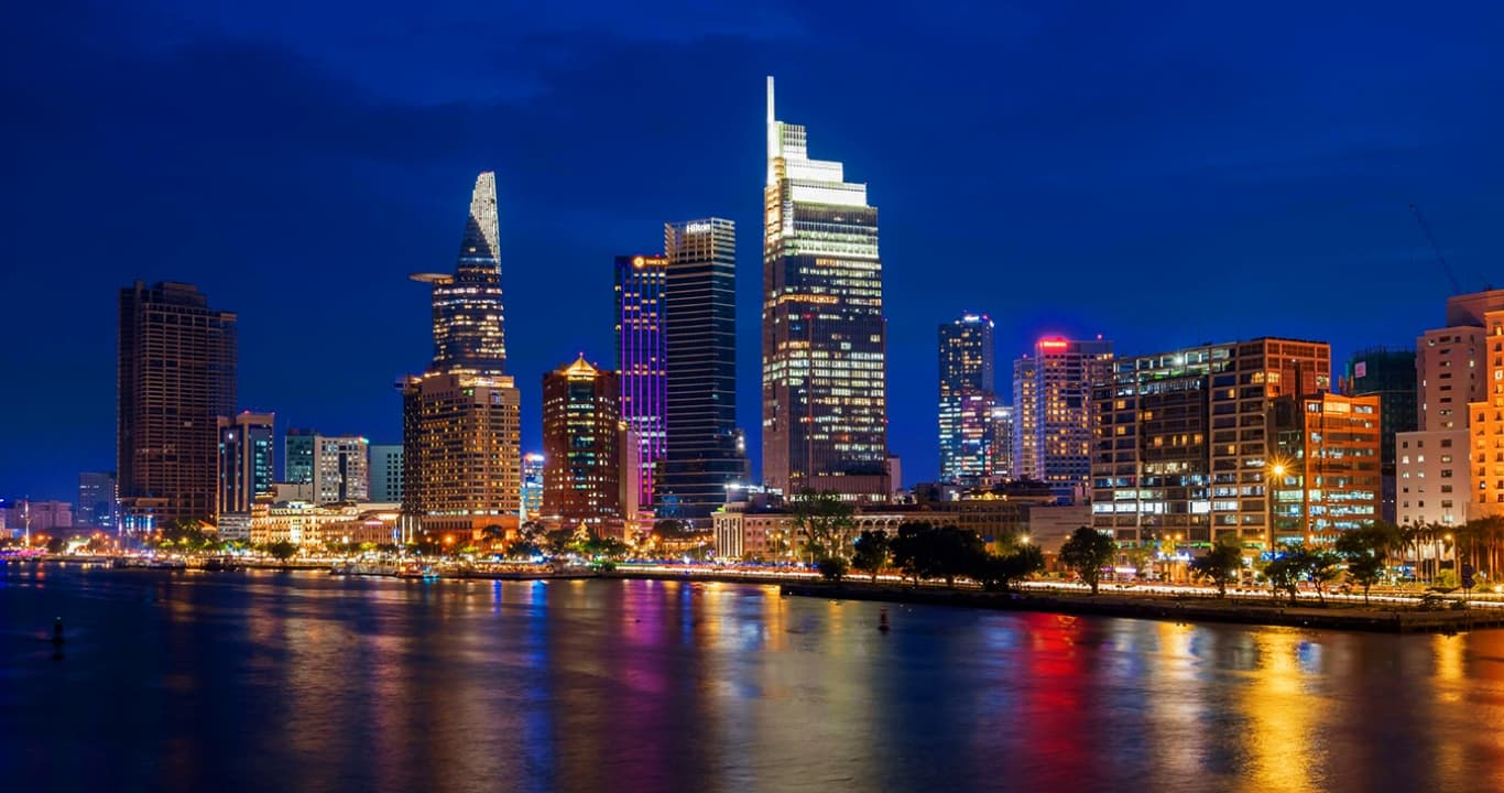 The Ho Chi Minh City skyline at night reflected in the Saigon River — Bitexco tower, Landmark 81, and the District 1 commercial core that anchor Vietnam's cost index