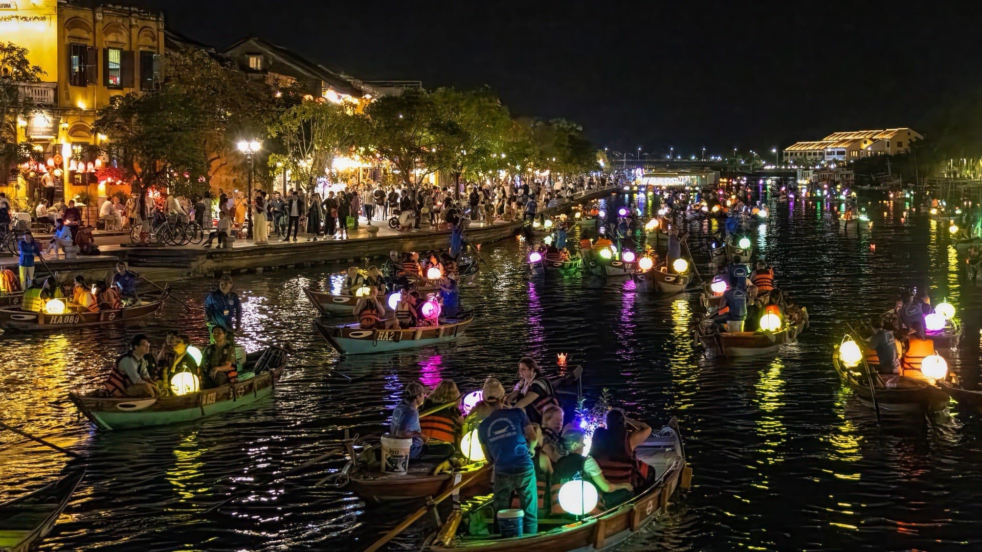 Rowing boats carrying glowing lanterns crowd the river beside Hoi An's lit-up old town waterfront at night, with people lining the promenade.