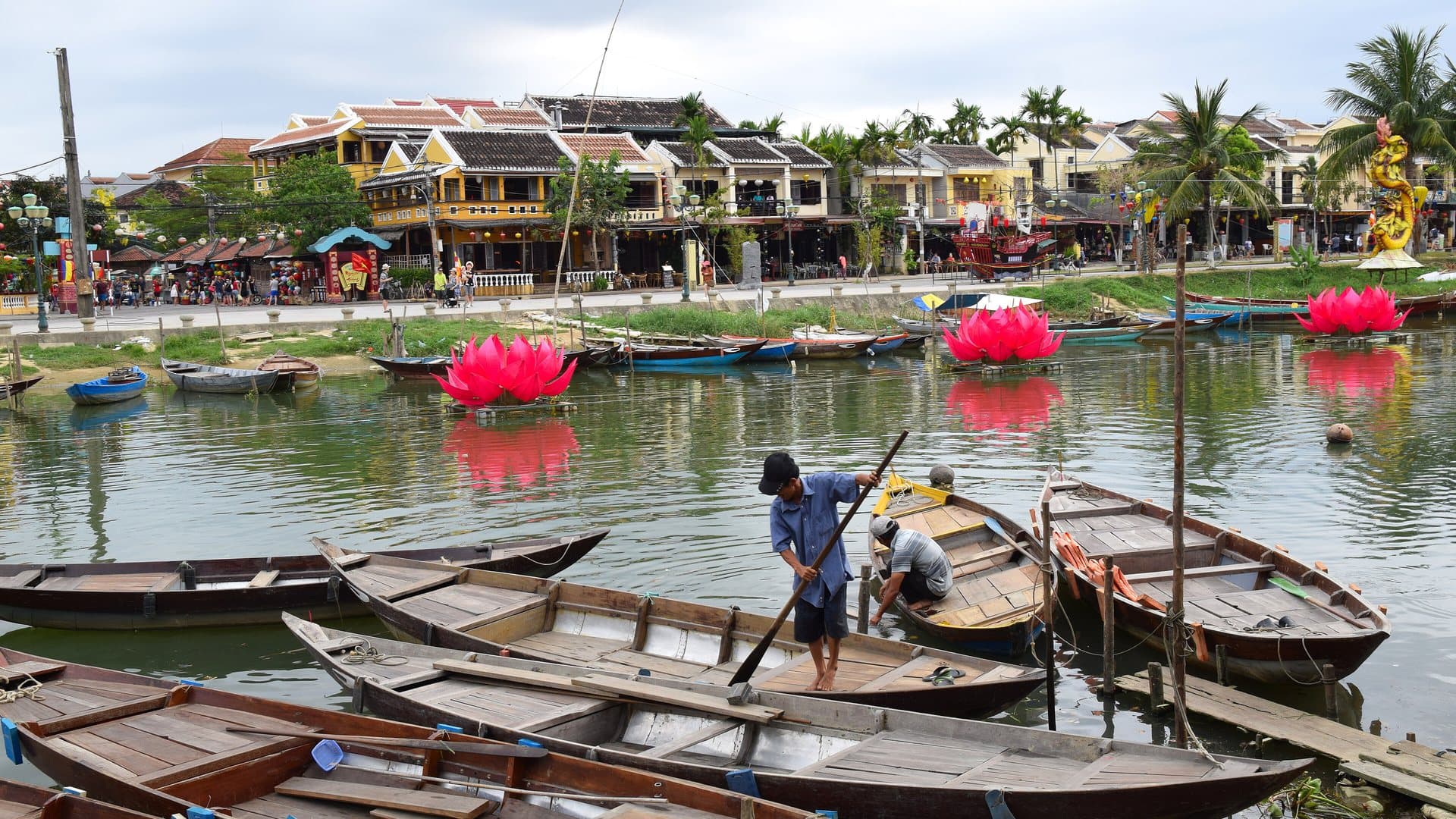 Wooden rowing boats moored on the Thu Bon River in Hoi An under an overcast sky, with pink lotus lanterns floating off the ochre riverfront houses
