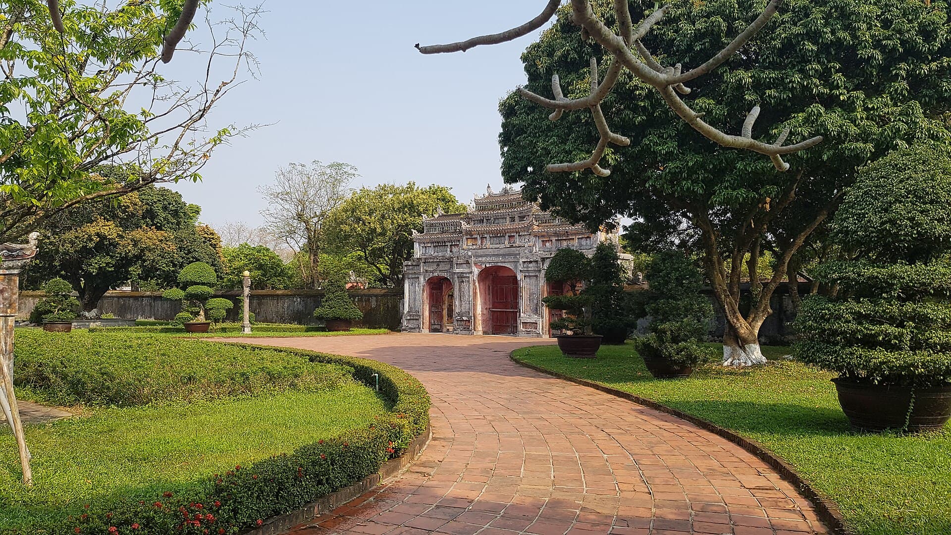 A wide, level brick path curving between clipped hedges, bonsai pots and shade trees toward the tiled ceremonial gate of Dien Tho Palace inside Hue's Imperial City
