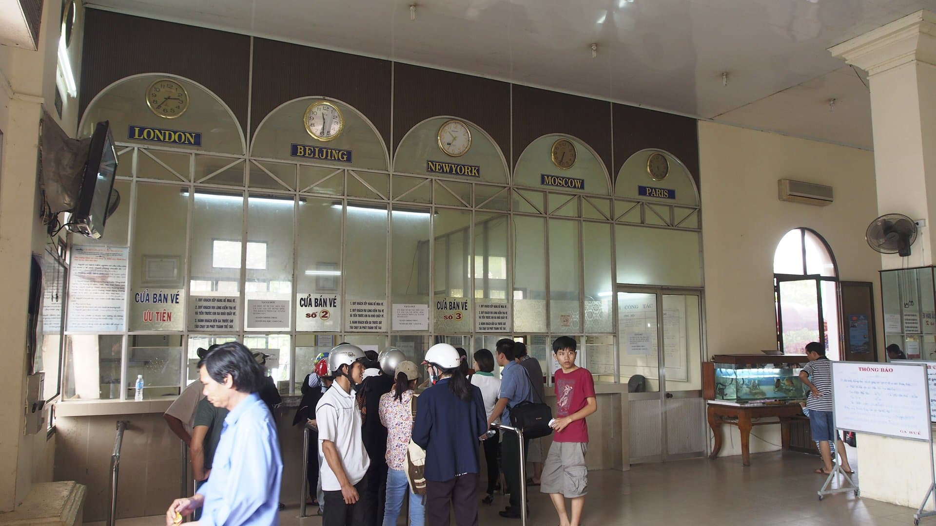 Passengers queuing at the ticket windows in the hall of Hue railway station, under arched windows and clocks