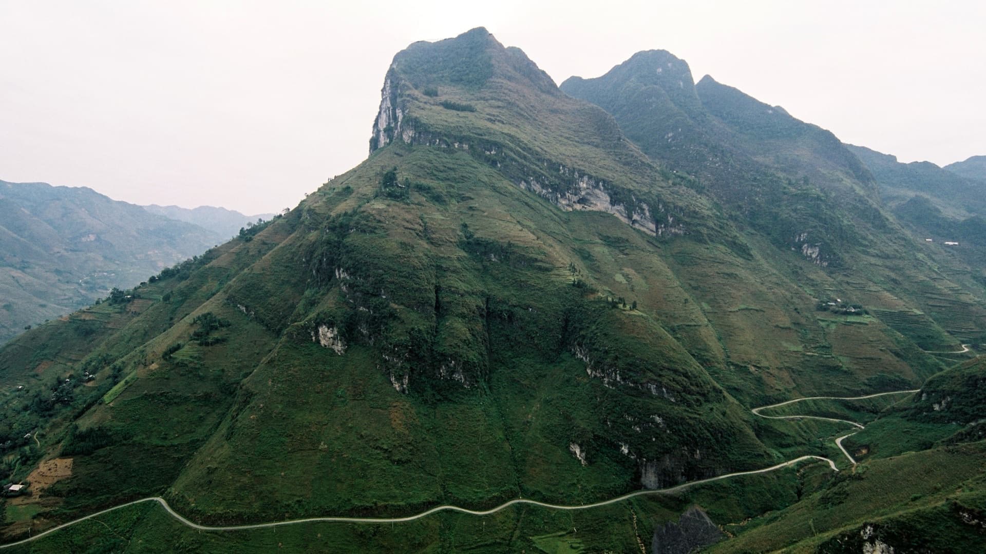 The Ma Pi Leng Pass road switchbacking across the lower slopes of a limestone peak in Ha Giang province, terraced fields and a few farmhouses in the valley below, under a flat overcast sky