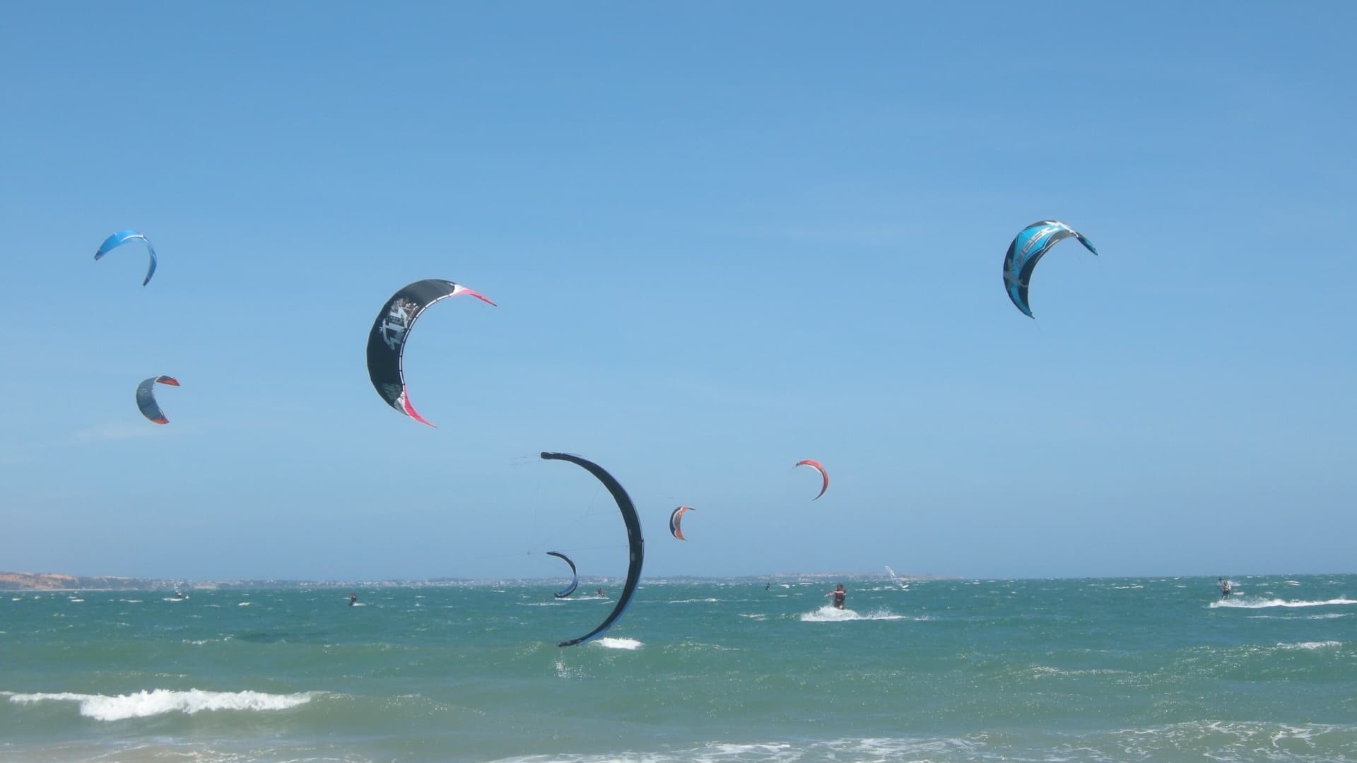 Kitesurfing kites fill a clear blue sky over the choppy turquoise sea off Mui Ne beach.