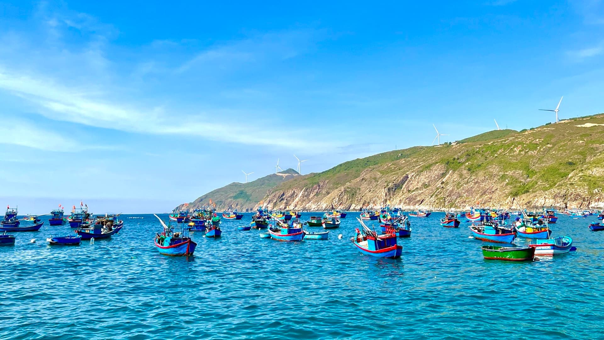Brightly painted fishing boats moored in the blue bay at Nhon Ly near Quy Nhon, below a rugged green headland lined with wind turbines.