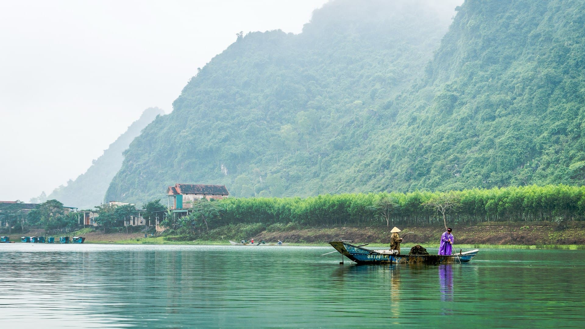 Two people work from a small wooden boat on the calm green Son River beneath a steep, forested limestone mountain wrapped in mist, with trees and a house along the far bank.