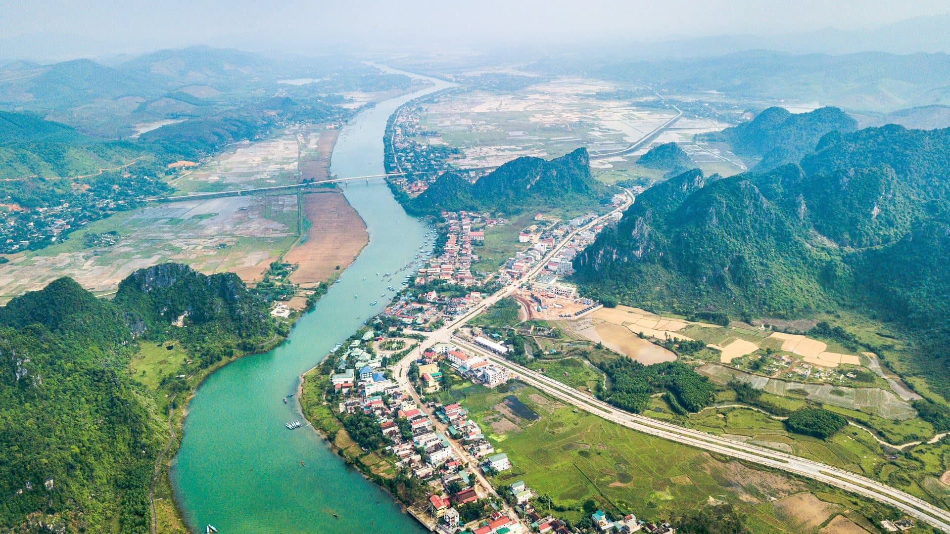 An aerial view shows the turquoise Son River winding past the houses of Phong Nha, with rice fields, a highway and forested limestone karsts fading into the haze.