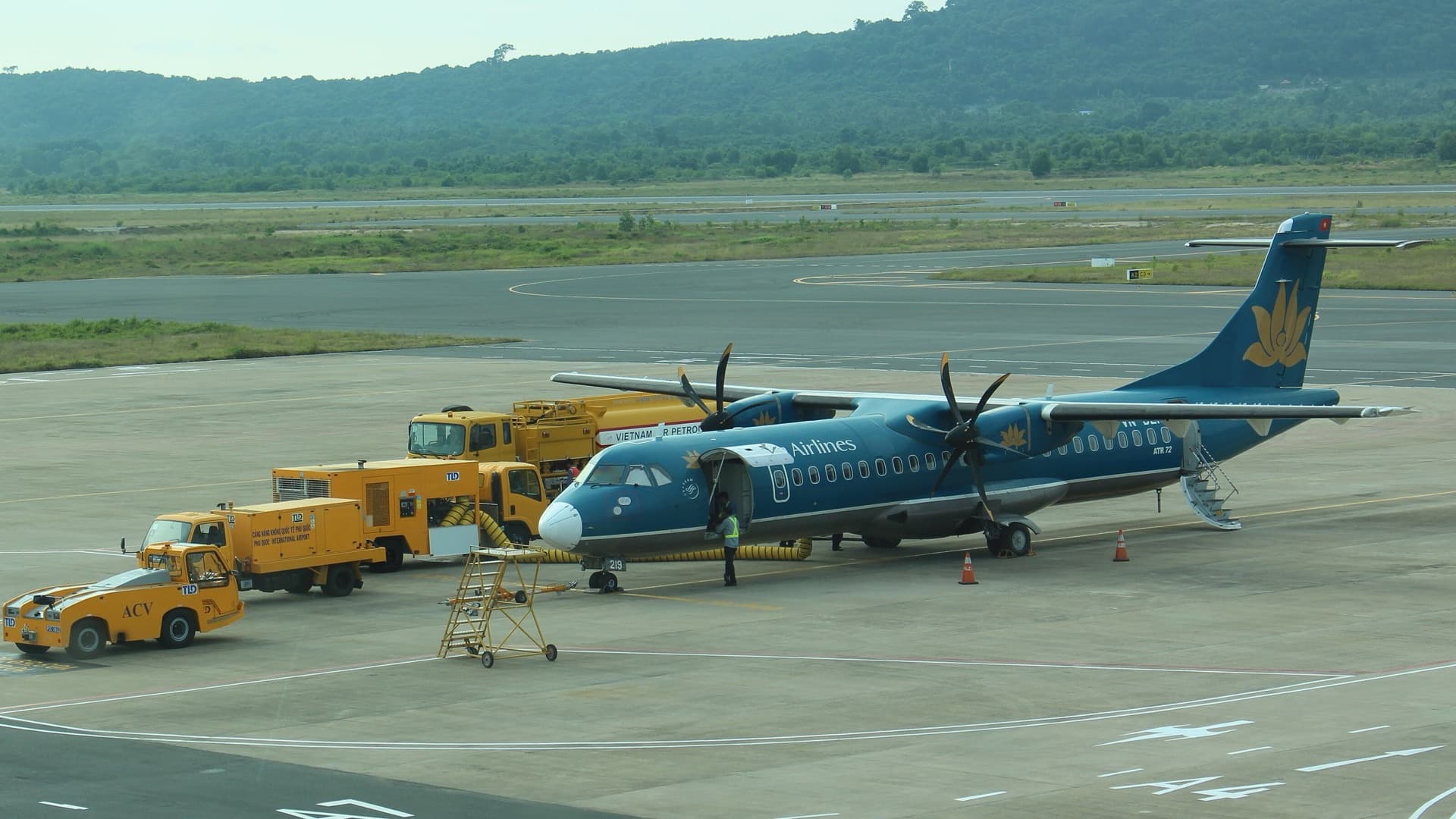 A Vietnam Airlines ATR 72 turboprop parked on the apron at Phu Quoc International Airport, with yellow service vehicles alongside and forested hills behind.