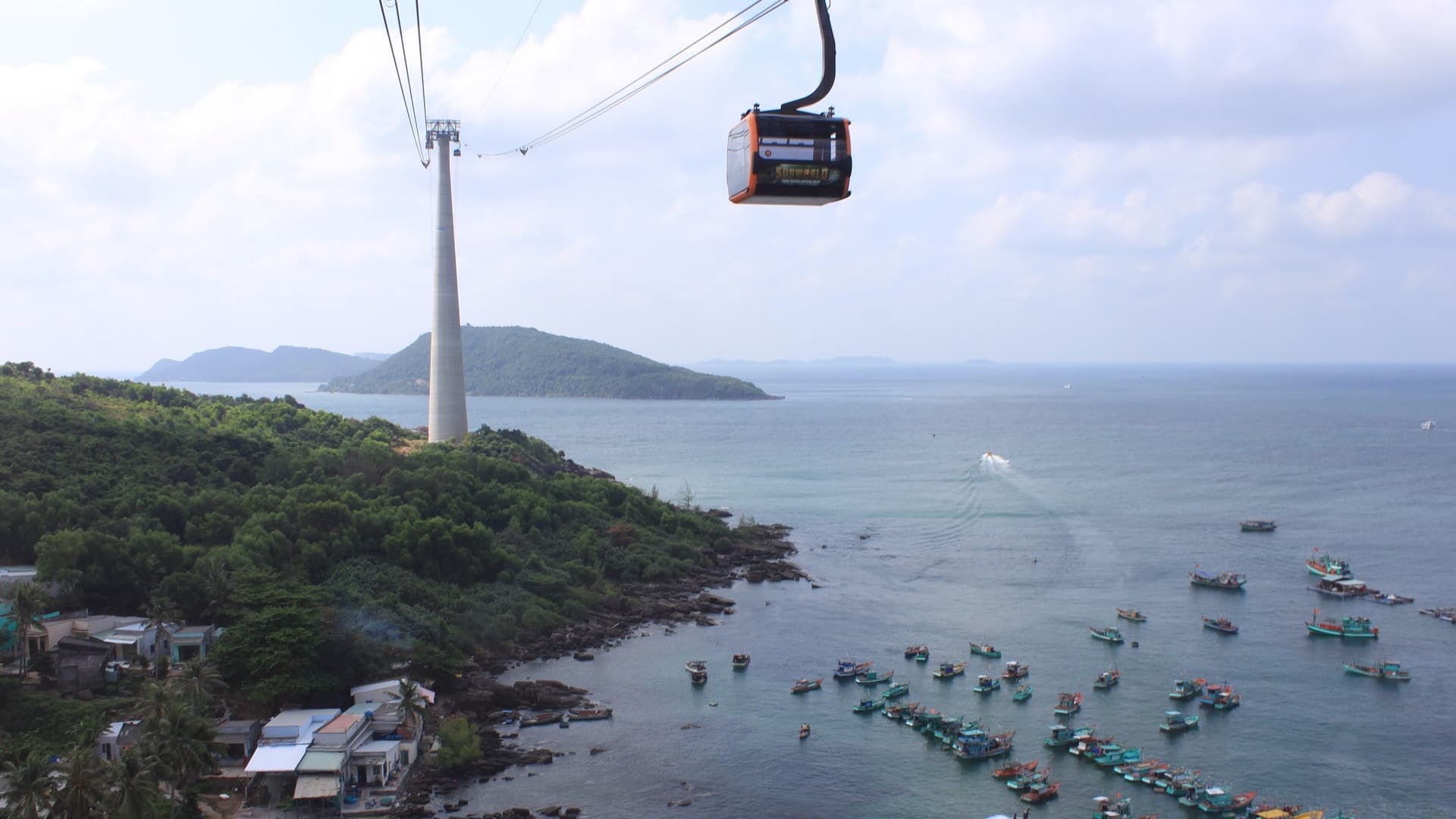A Hon Thom cable car cabin passes above the forested coast of An Thoi on Phu Quoc, with a tall pylon, fishing boats and offshore islands below.