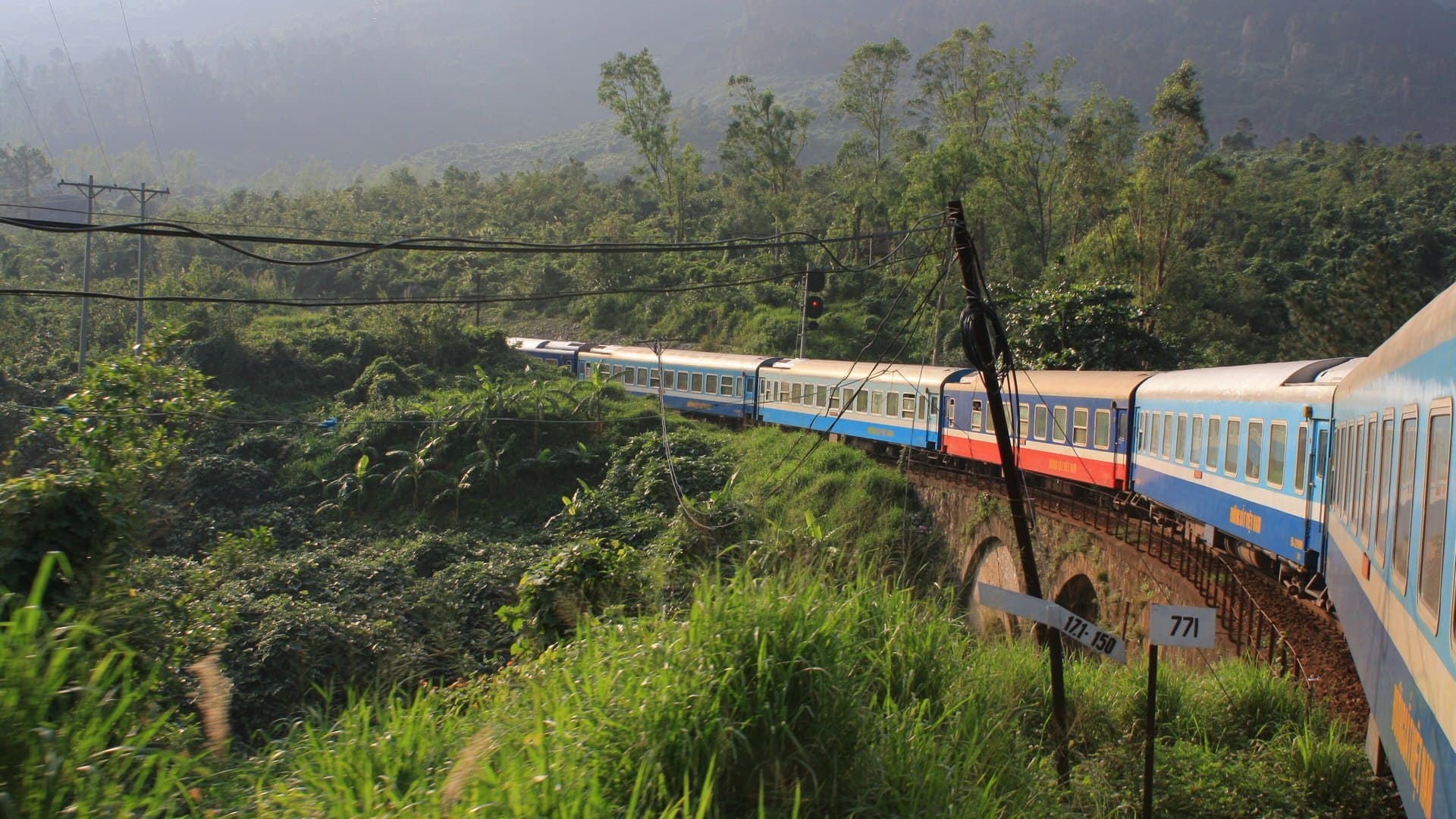 A blue-and-white Vietnam Railways passenger train curves across a brick arch viaduct through a lush green hillside, seen from a window of the same train.