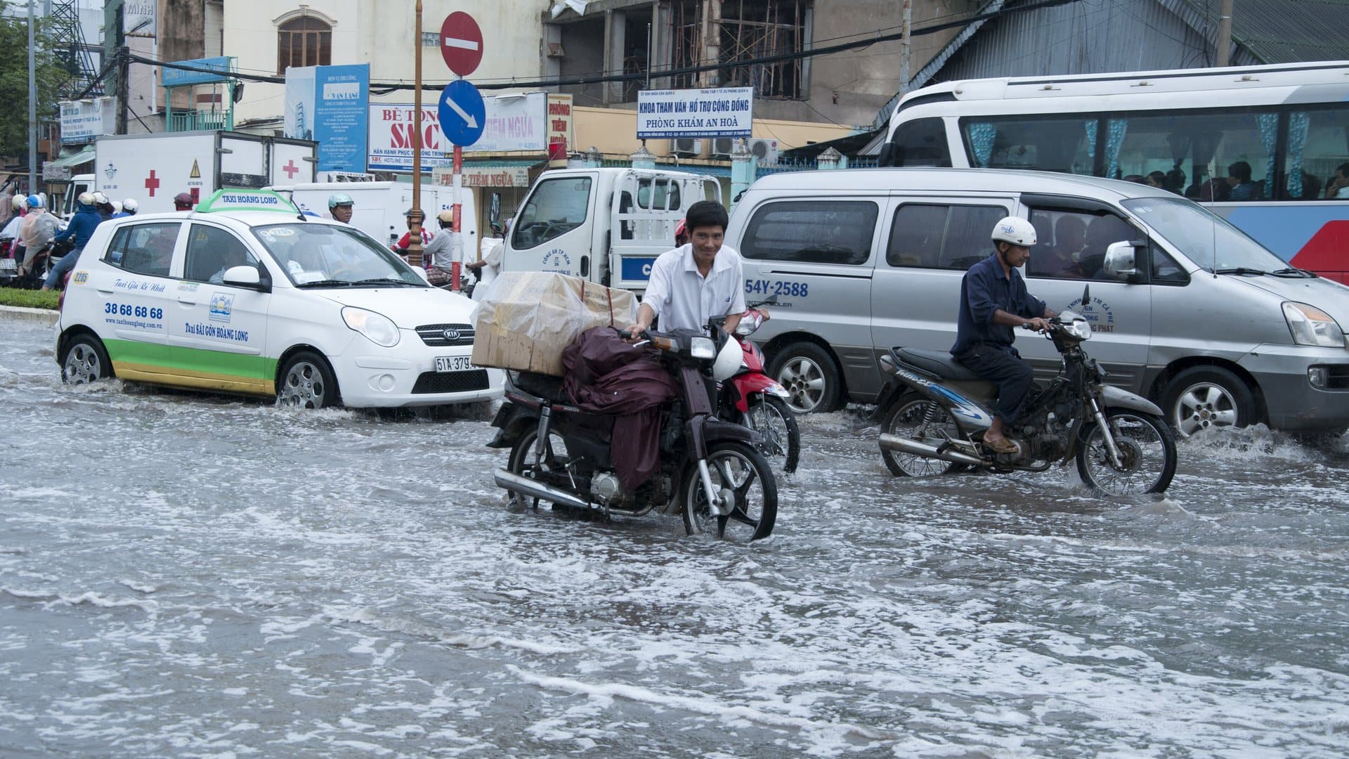 Motorbikes and a taxi splashing through a rain-flooded street in Ho Chi Minh City during the rainy season, a familiar sight on wet-season afternoons