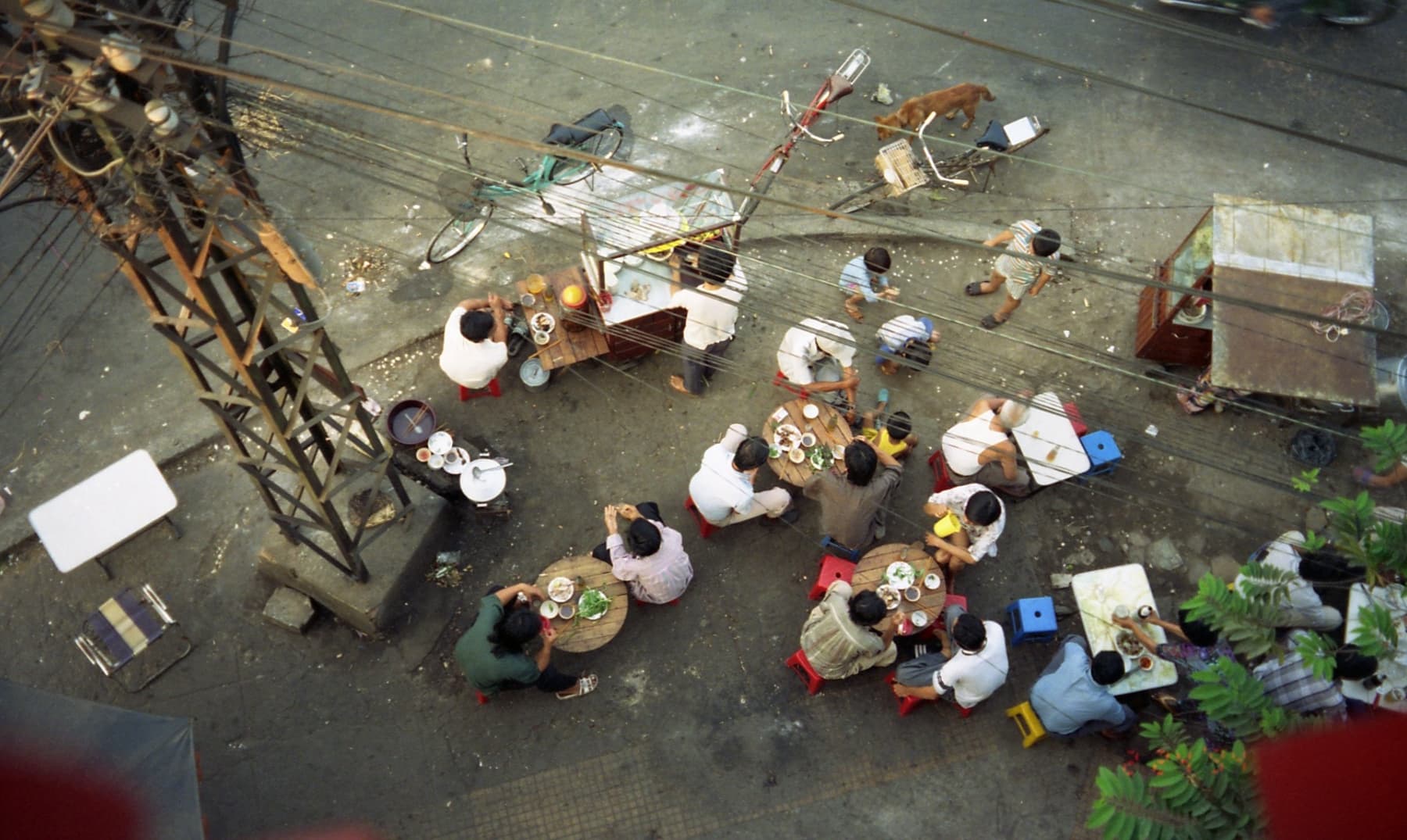 Overhead view of a Saigon street corner with multiple groups eating Vietnamese street food at low plastic tables and stools — the everyday architecture of Vietnamese dining culture
