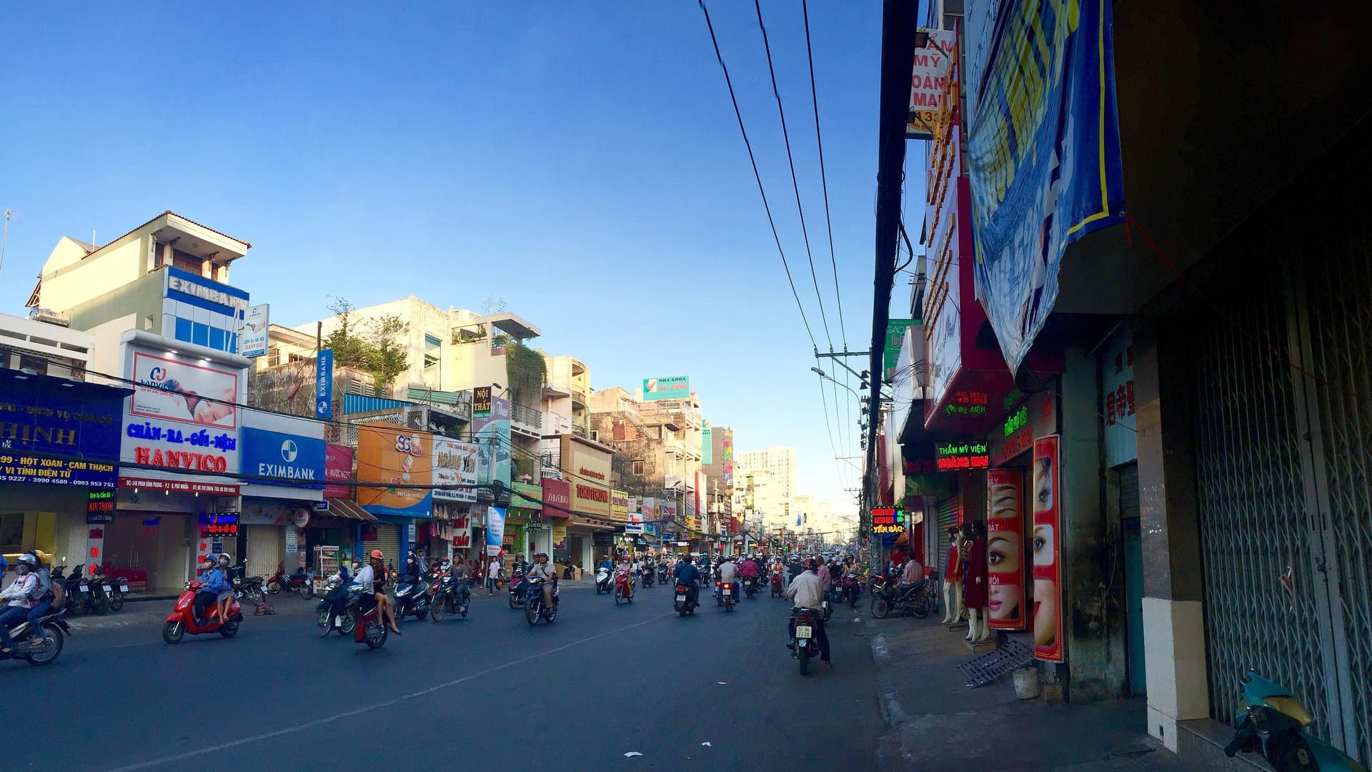 A busy Ho Chi Minh City street lined with shopfronts, with bundles of overhead power lines on the right and motorbikes in the traffic
