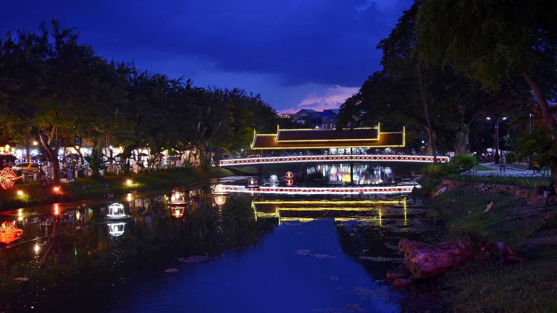 An illuminated covered footbridge and colourful lights reflect in the Siem Reap River at dusk, framed by tall riverside trees.