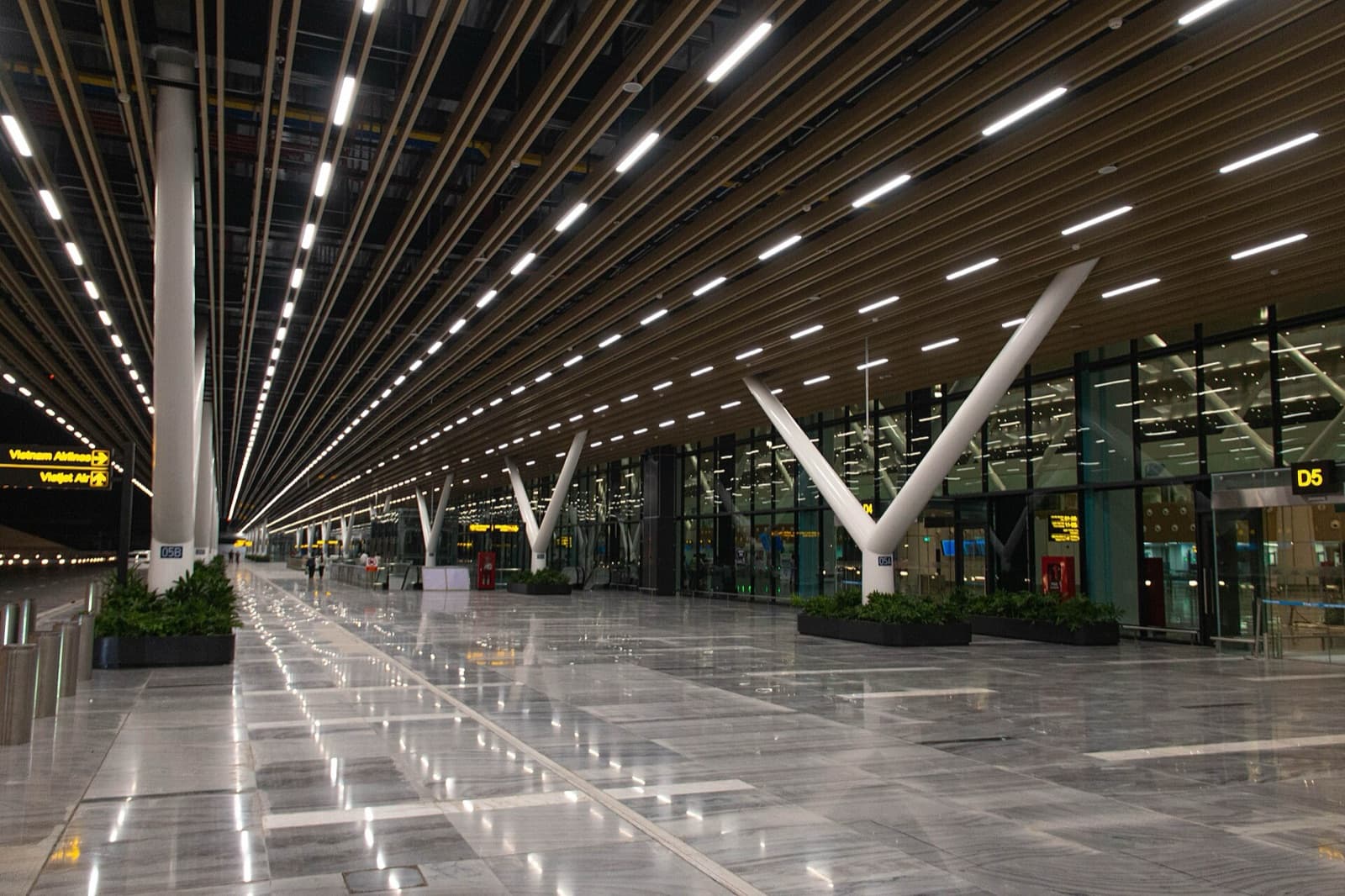 The near-empty departures concourse of Terminal 3 at Tan Son Nhat International Airport in Ho Chi Minh City, its timber-slat ceiling and white Y-columns running past Vietnam Airlines and Vietjet Air wayfinding signs