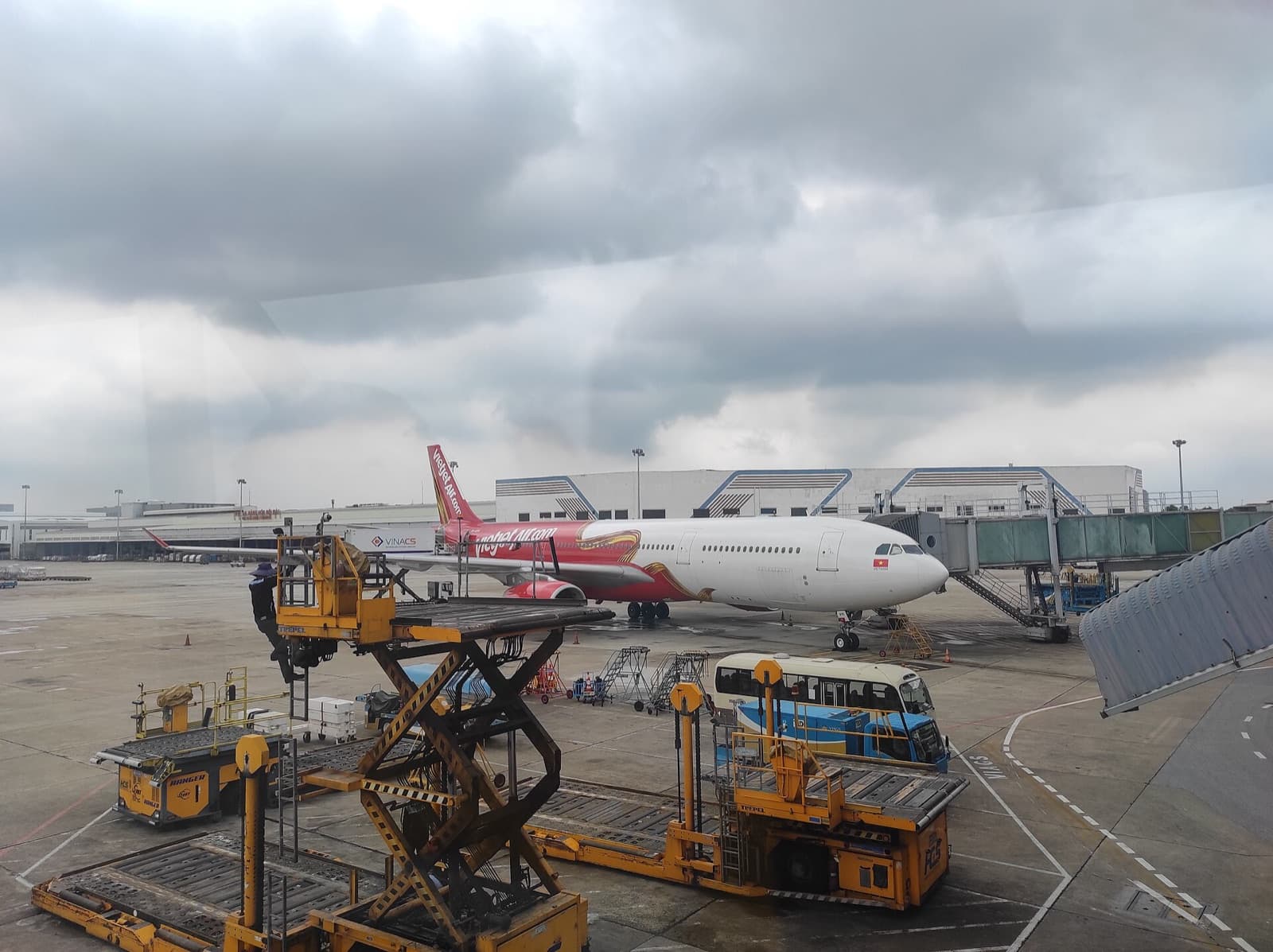 A VietJet Air Airbus A330 parked at a jet bridge at Noi Bai International Airport in Hanoi under heavy monsoon cloud, with baggage loaders and a crew bus on the apron