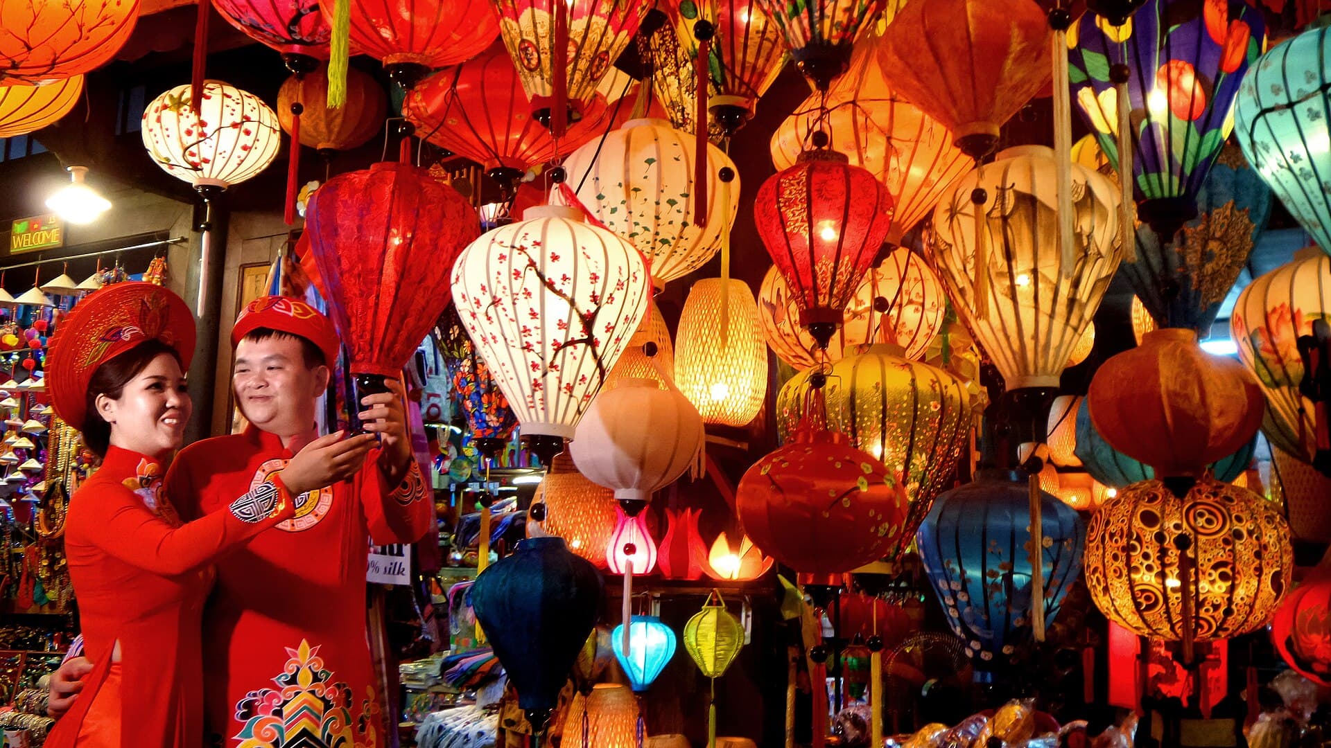 A Vietnamese couple in red ao dai and khan dong headwear smiling together beneath dozens of glowing silk lanterns inside a Hoi An lantern shop