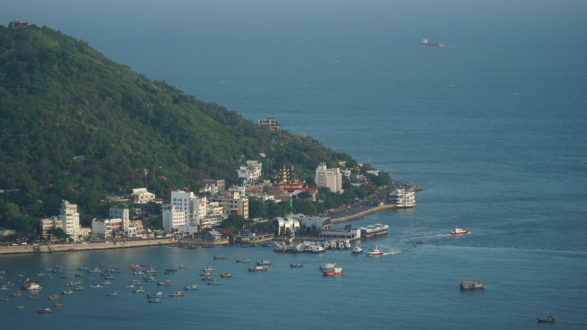 A green wooded headland slopes down to Vung Tau's Front Beach waterfront, where a tiered pagoda, white buildings and a ferry pier overlook dozens of fishing boats moored in the bay.