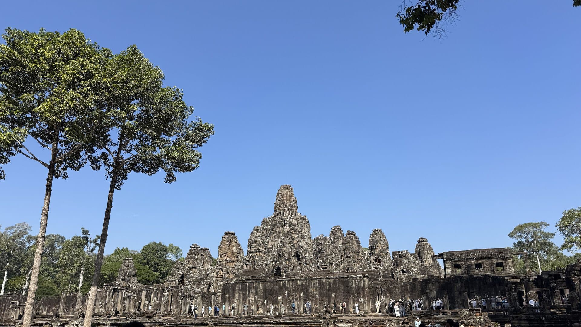 The stone face towers of the Bayon temple at Angkor Thom rising against a cloudless dry-season sky, framed by tall trees, near Siem Reap in Cambodia
