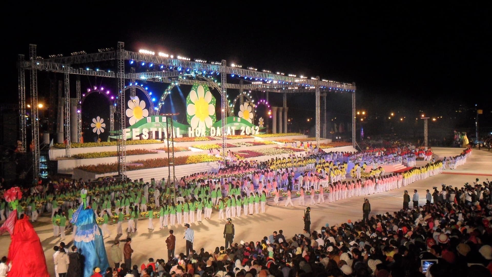 Opening-night stage with flower-covered tiers, hundreds of performers in coloured uniforms and a packed standing crowd at the 2012 Da Lat Flower Festival