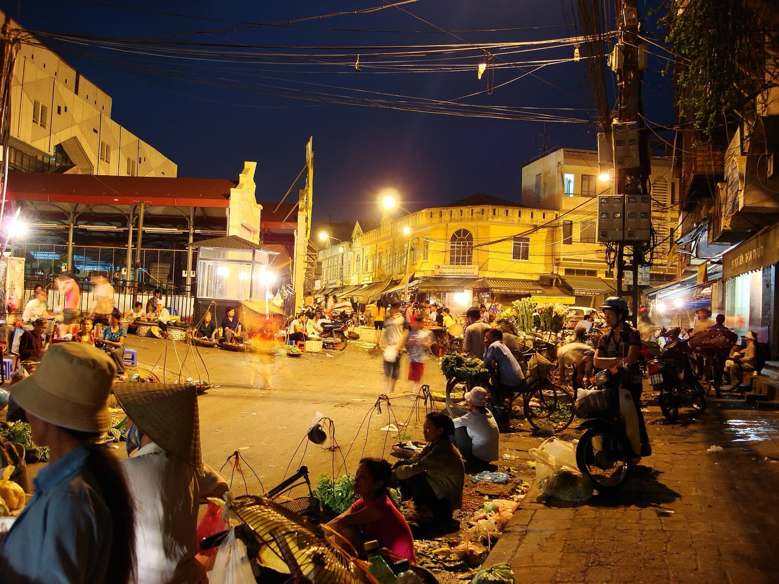Vendors in conical hats selling greens and fruit from shoulder-pole baskets set on the ground at the night street market beside Dong Xuan Market in Hanoi's Old Quarter, lit by street lamps at dusk