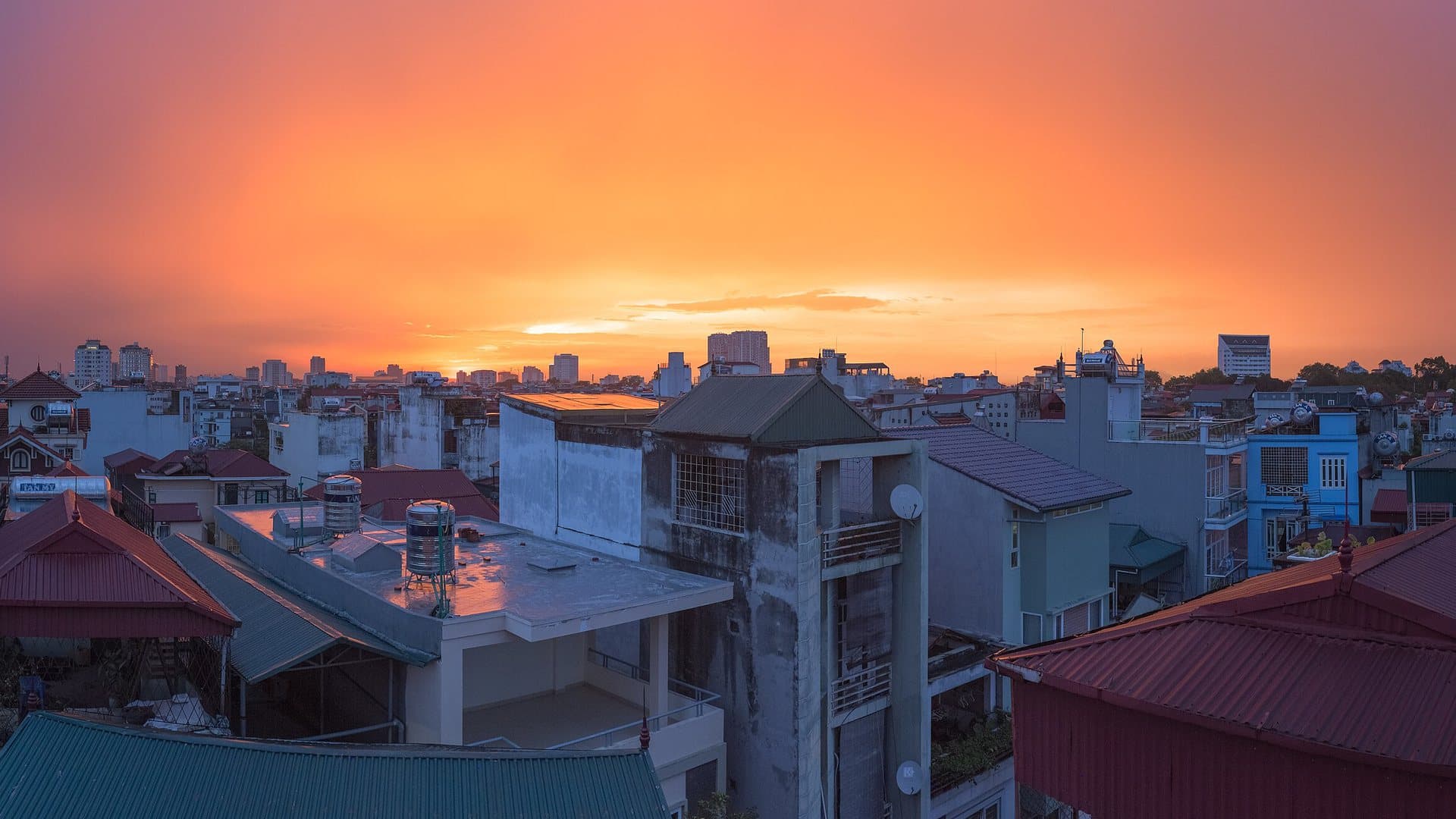 An orange sunset sky clearing over Hanoi's rooftops and water tanks after a rainstorm, with the city's towers on the horizon