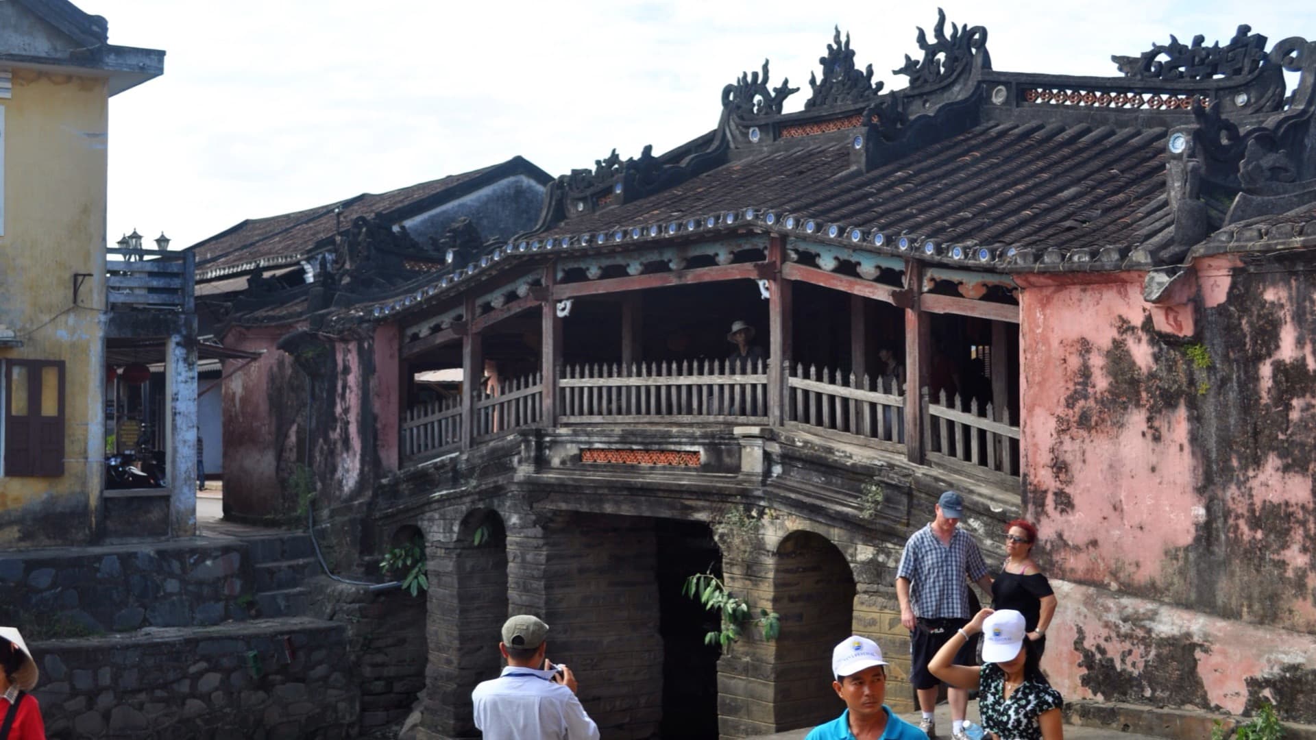 The Japanese Covered Bridge in Hoi An seen side-on in daylight, its dark tiled roof and timber balustrade set between weathered pink-plaster end walls, with a few visitors standing below