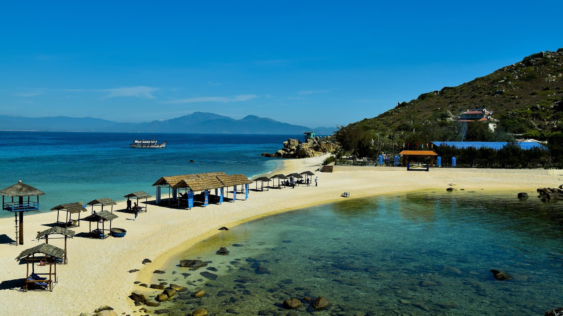 A crescent of white sand, thatched beach umbrellas and clear turquoise water on Hon Noi island in Nha Trang Bay under a blue dry-season sky