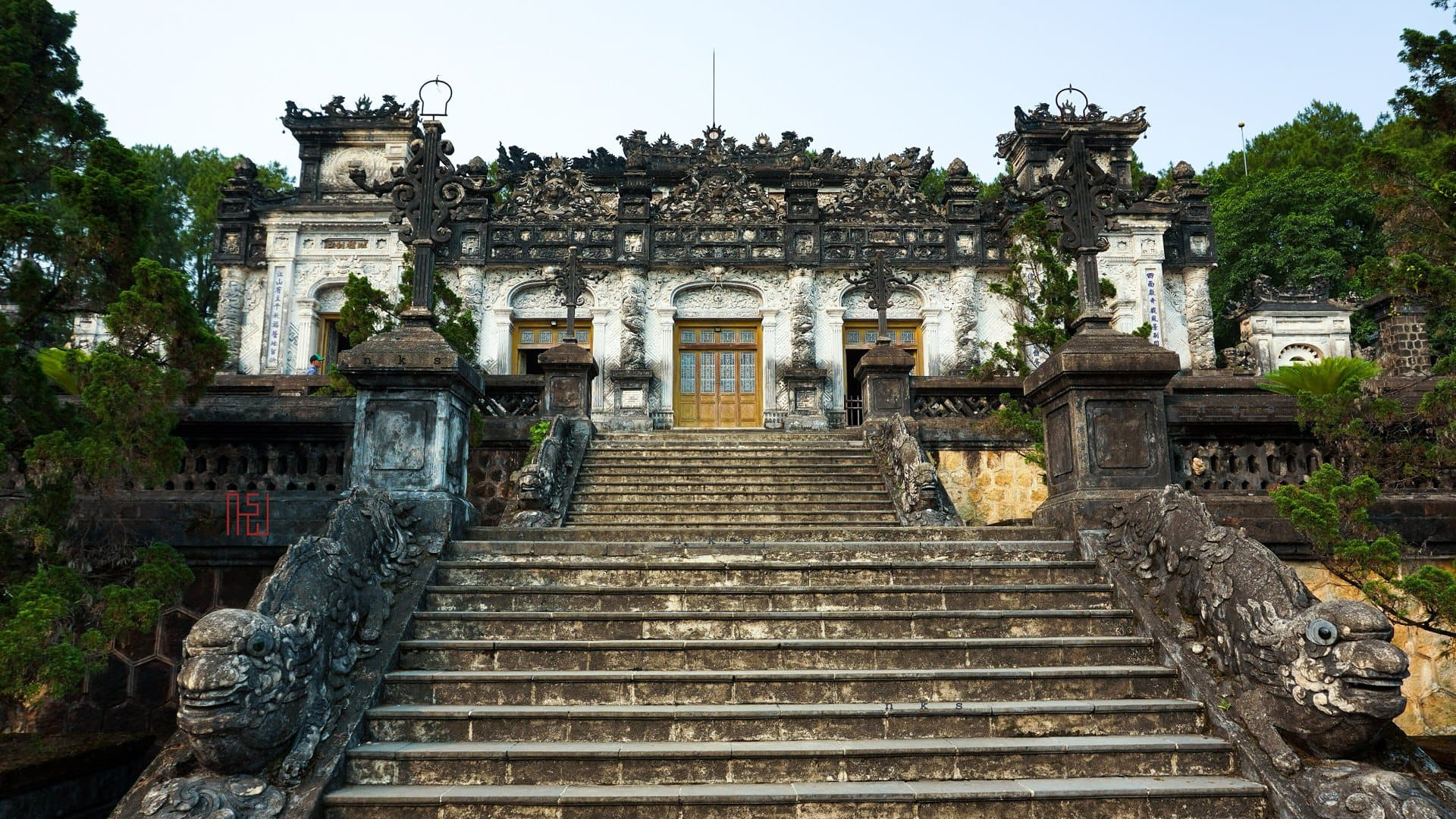 A broad stone staircase flanked by dragon balustrades climbs to the ornate grey-and-white facade of Khai Dinh's tomb in Hue, framed by trees and a pale sky.