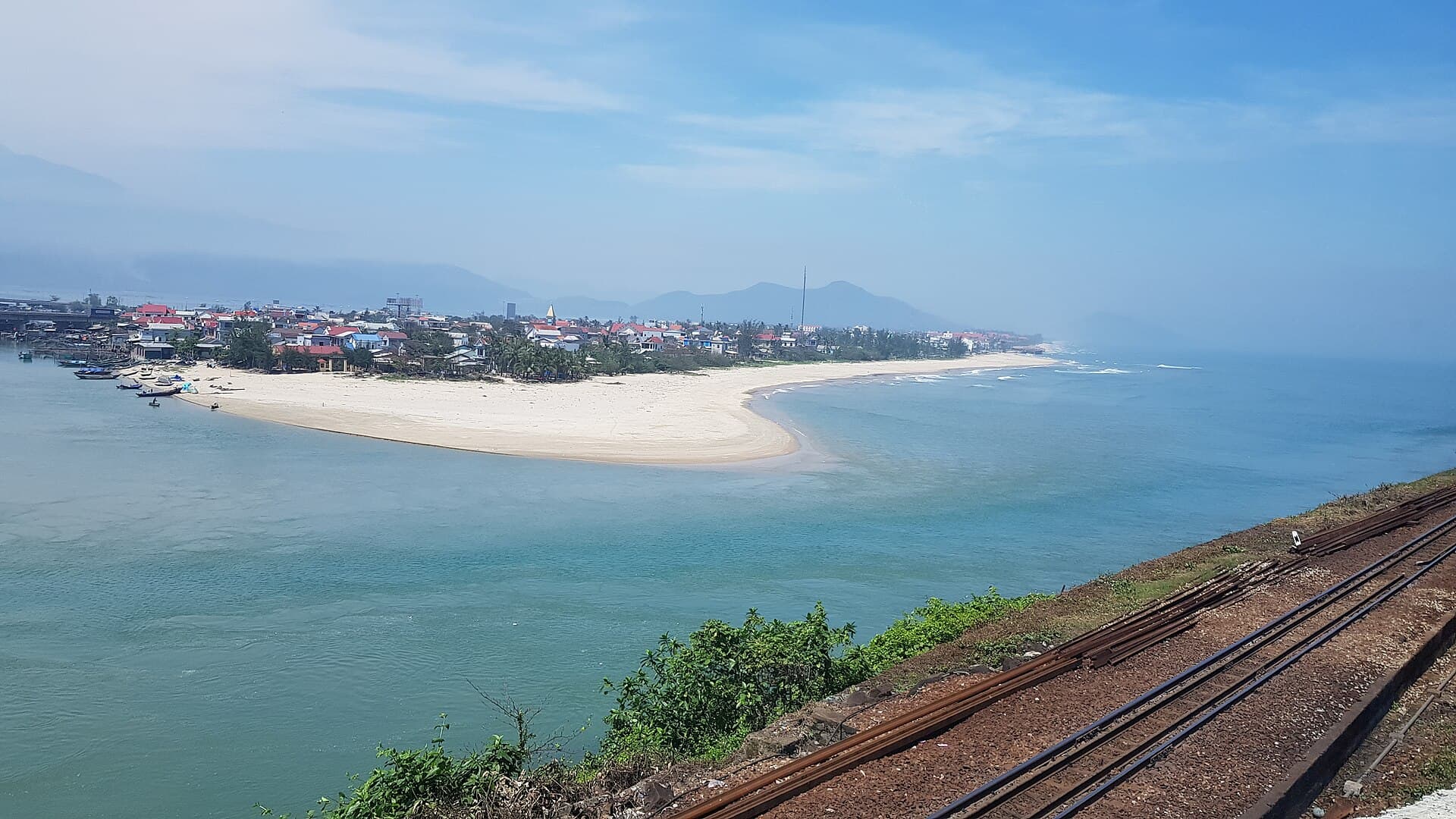 The white sand spit at Lang Co on Vietnam's central coast, where the lagoon meets the sea beside a red-roofed fishing village, with the Reunification Express tracks running along the hillside in the foreground