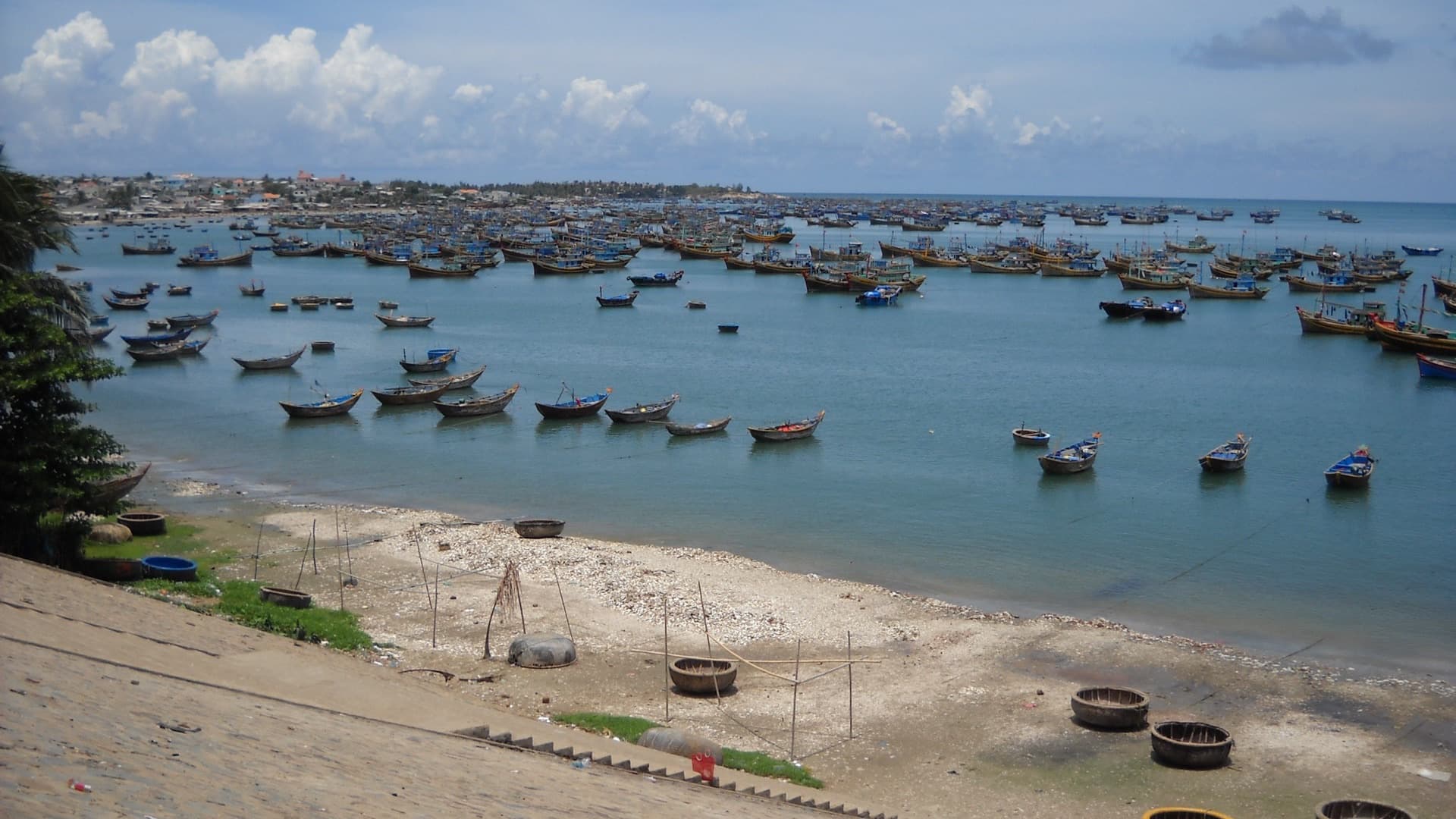 Dozens of fishing boats and round basket boats anchored in the bay off Mui Ne fishing village, seen from above the beach.
