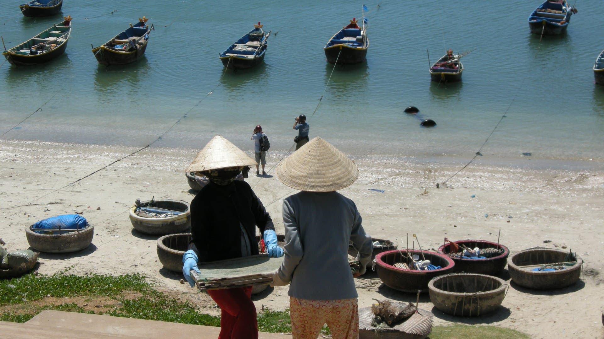 Two women in conical hats carrying a tray across the beach at Mui Ne fishing harbour, round coracle boats resting on the sand behind them and blue wooden fishing boats moored in the shallows