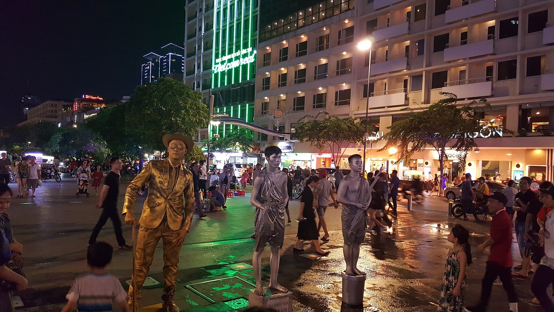 Crowds filling Nguyen Hue walking street in Ho Chi Minh City at night, with gold and silver living-statue performers in the foreground and lit tower blocks behind