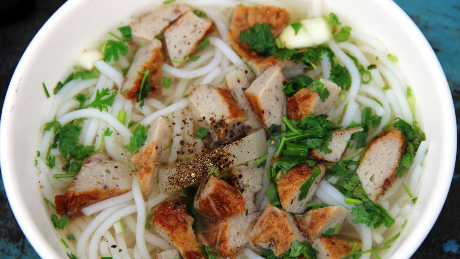 A white bowl of bun cha ca holds round rice noodles, sliced golden fish cake, chopped herbs and a sprinkle of black pepper in clear broth.