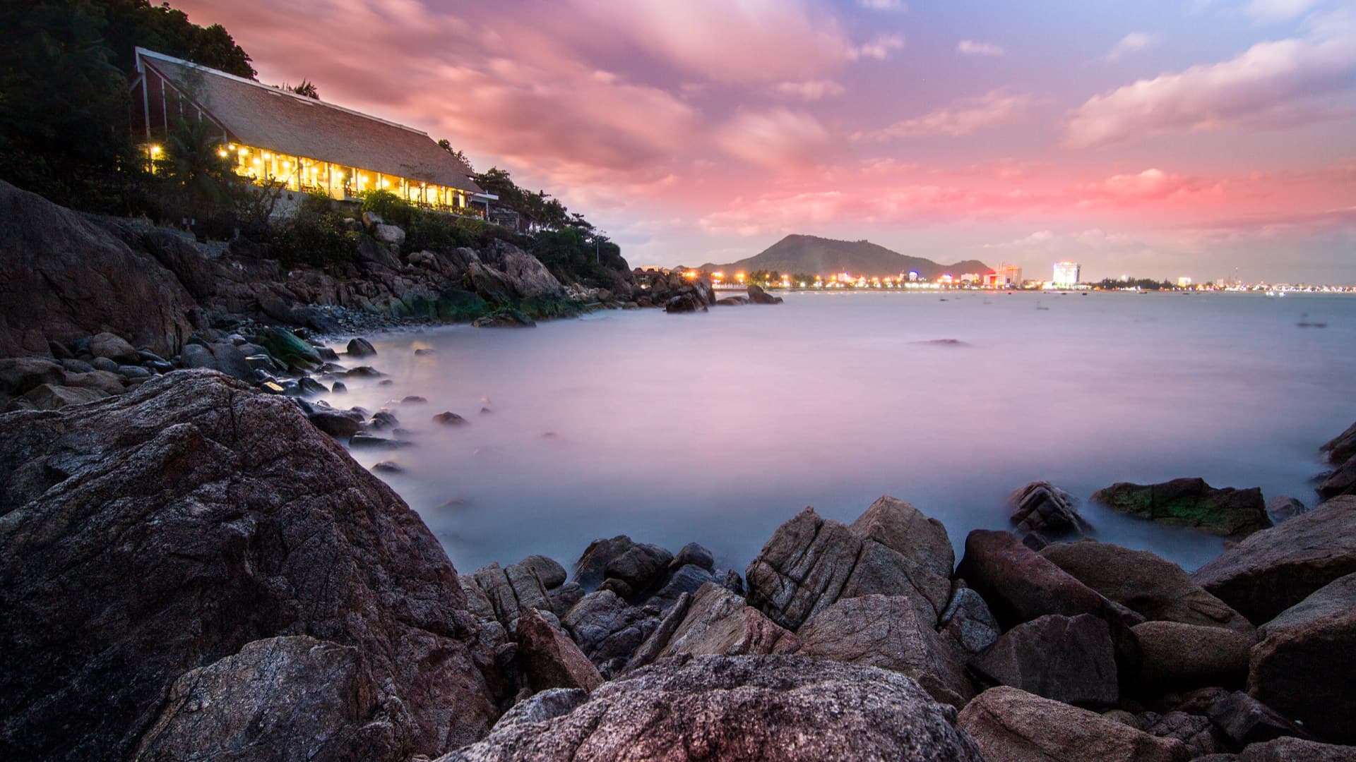 At dusk, misty water washes around the boulders of the Ghenh Rang shore beneath a lit hillside building, with Quy Nhon's city lights and a mountain across the bay under a pink sky.