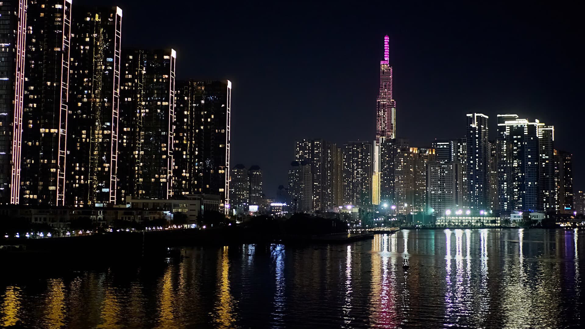 Landmark 81 lit pink above a wall of riverside apartment towers, their lights reflected in the black water of the Saigon River at night